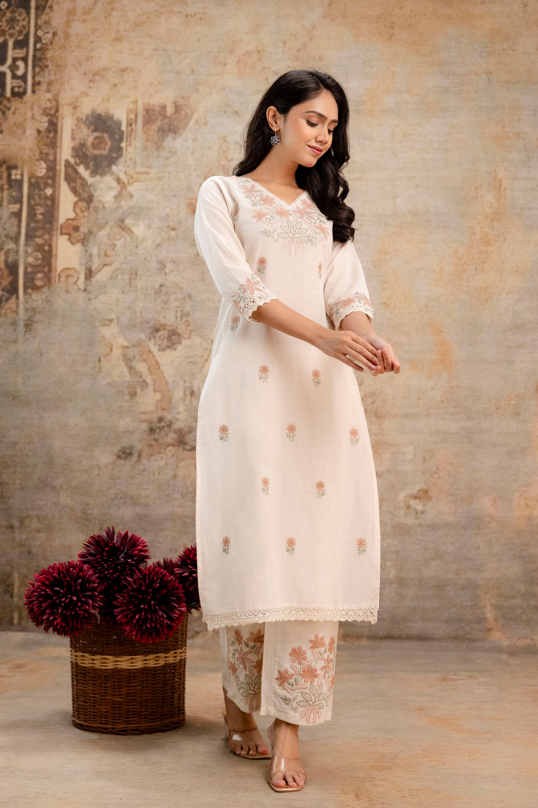 A woman wearing a Tanauri cotton kurti set in a soft off-white/beige tone.
The kurti features delicate peach and green floral embroidery scattered across the front and along the neckline and sleeve edges.
The neckline is V-shaped with intricate lace trimming, enhancing the elegant and traditional appeal.
The sleeves are three-quarter length, adorned with matching embroidery and lace detailing.
She pairs it with straight-fit cotton pants featuring heavier floral embroidery at the bottom hem, echoing the 