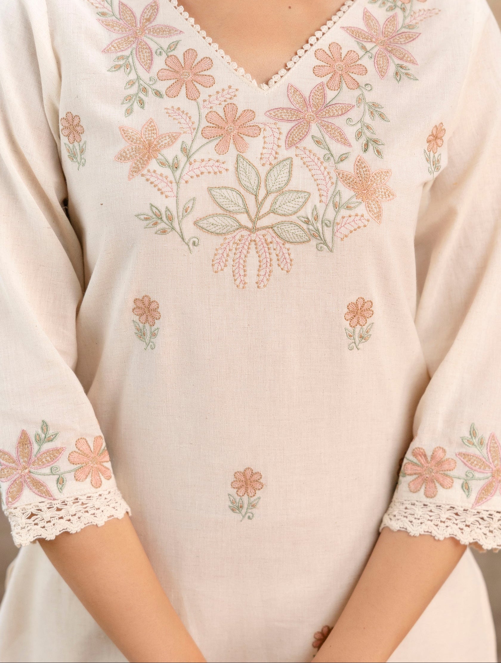 Close-up view of a Tanauri cotton kurti in a soft off-white shade.

Focus on the intricate floral embroidery around the V-neckline and chest area.

The embroidery features pastel peach, pink, and green floral motifs, including daisies and leafy patterns.

Delicate lace trim outlines the neckline and sleeve cuffs, adding a feminine and handcrafted touch.

Fabric texture is visible, showcasing breathable, soft cotton ideal for summer wear.

Embroidery on sleeves and bodice is symmetrically placed, enhancing 