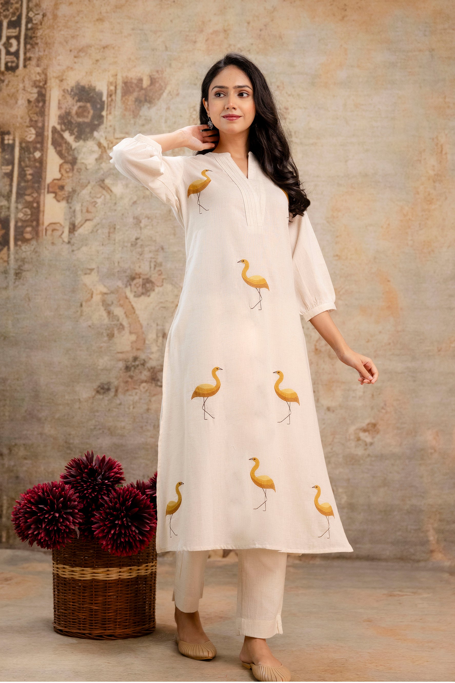 Elegant ivory kurti set with mustard yellow crane/bird motifs printed symmetrically across the front.

Features a high V-slit neckline with delicate pintuck detailing at the yoke.

Designed with three-quarter balloon sleeves gathered at the cuffs for a breezy silhouette.

Made from breathable, lightweight fabric – ideal for warm-weather outings.

Paired with coordinated ivory ankle-length straight pants.

Keywords: ivory bird print kurta set, printed cotton kurti with pants, summer kurta design for women