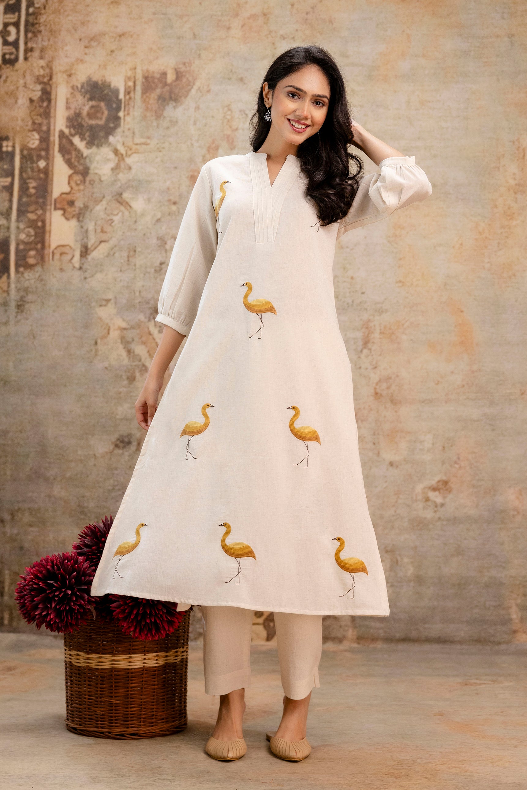 Ivory A-line kurta set featuring vivid mustard yellow crane bird prints evenly distributed across the front.

The kurta has a subtle mandarin V-neckline with fine pintuck stitching on the yoke.

Styled with three-quarter sleeves ending in a clean hem for an elegant silhouette.

Made from breathable cotton or cotton-linen blend for a light, summery appeal.

Paired with straight-fit ivory trousers that complement the kurta's neutral tone.
Keywords: mustard crane print kurti, ivory cotton kurta set, summer kur