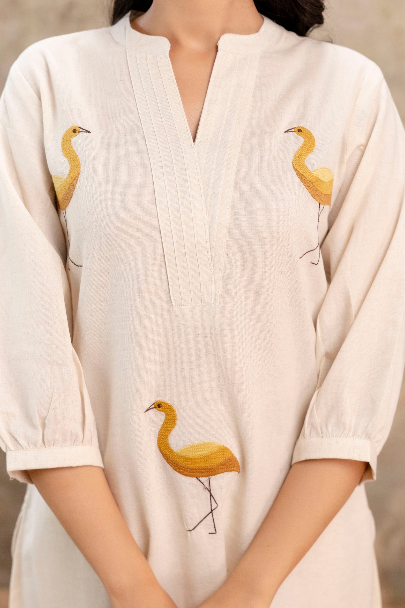 Close-up of an ivory kurta with mustard crane embroidery on front and sleeves.

Features a Mandarin collar with a deep V-slit and pintuck detail.

Made from soft cotton fabric with a subtle crinkled texture.

3/4th sleeves with lightly gathered cuffs.

Highlights minimalist ethnic style with artisanal embroidery.

Keywords: embroidered kurta, crane motif, ethnic wear, women's cotton kurti.