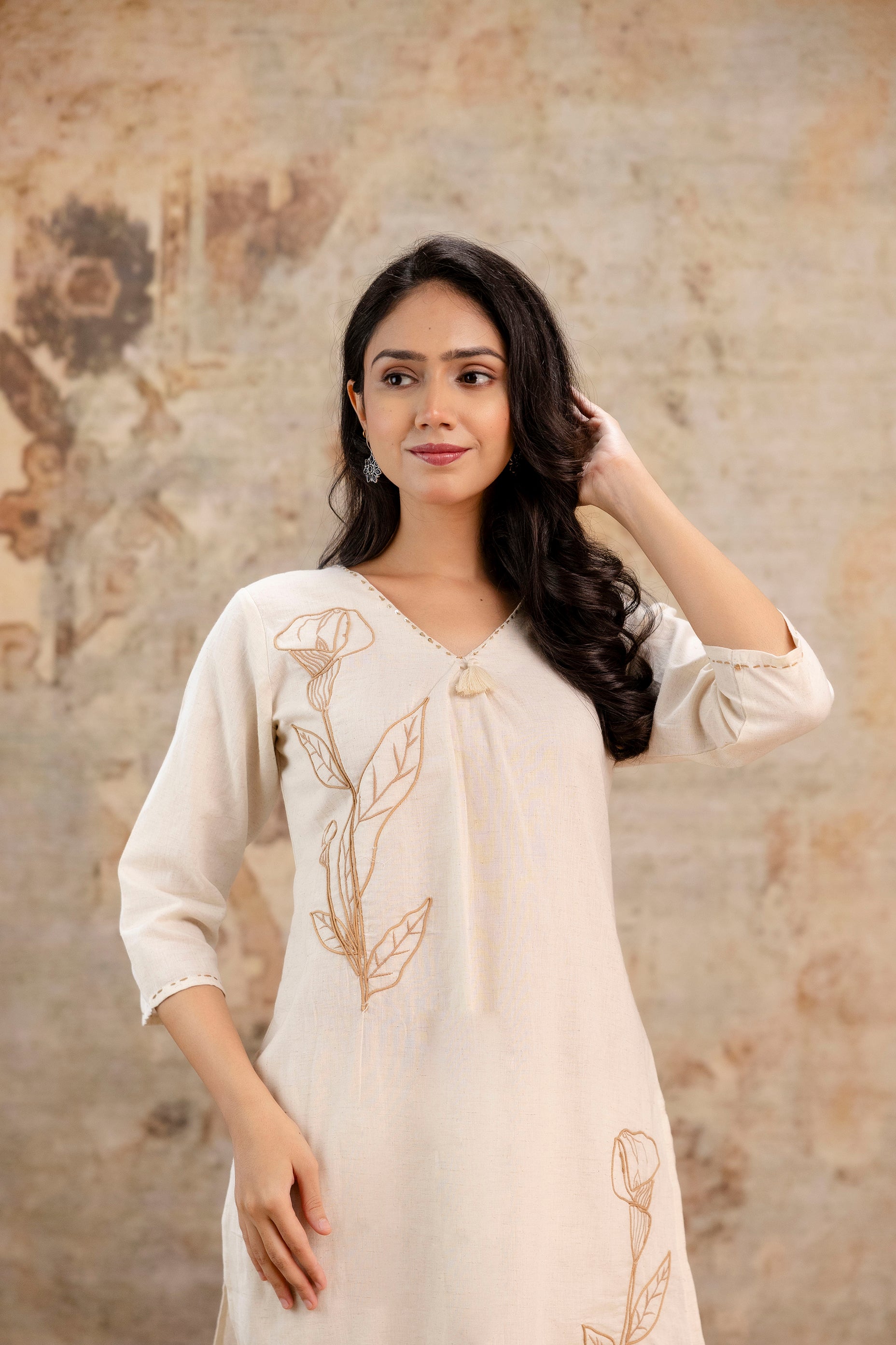 Woman modeling a cream-colored kurti with brown floral embroidery.

Embroidery features a large stitched flower with leaves on the left side of chest and thigh.

Kurti has a V-neckline with minimal border detailing and 3/4th sleeves.

Model has long, curled hair swept to one side, lightly touching it with her right hand.

Subtle earthy-toned background enhancing the natural and elegant vibe of the outfit
