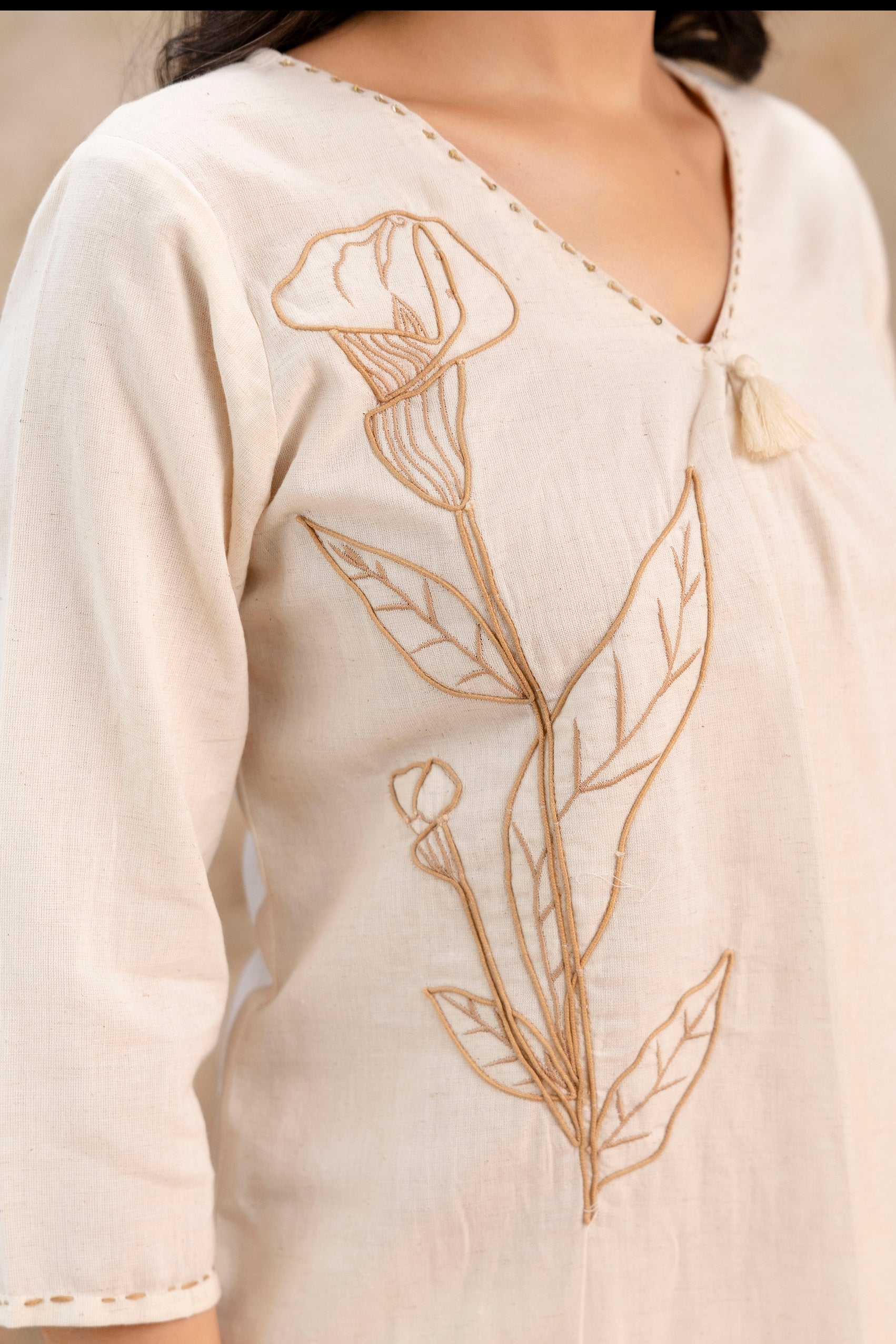 Off-white kurti set with full-length embroidered floral design in beige thread

Features a deep V-neckline with delicate stitched piping and front tassel

Three-quarter sleeves with matching embroidery on the left side

Paired with straight-fit off-white pants, also embroidered near the hem

Made from soft, breathable fabric ideal for casual and semi-formal occasions

Model styled with minimal accessories for a clean, elegant look
