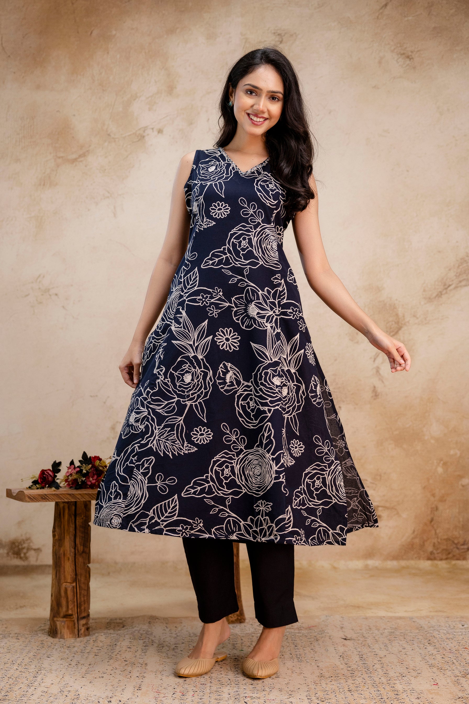 Woman wearing a sleeveless navy blue A-line kurti with bold white floral print.

Kurti features a flattering V-neckline and flared silhouette for elegant movement.

All-over white rose and botanical pattern creates a high-contrast statement look.

Paired with straight black pants for a balanced, contemporary ethnic outfit.

Styled with minimal accessories and natural makeup, perfect for casual or semi-formal wear.

Beige juttis add a subtle traditional touch to the modern printed ensemble.