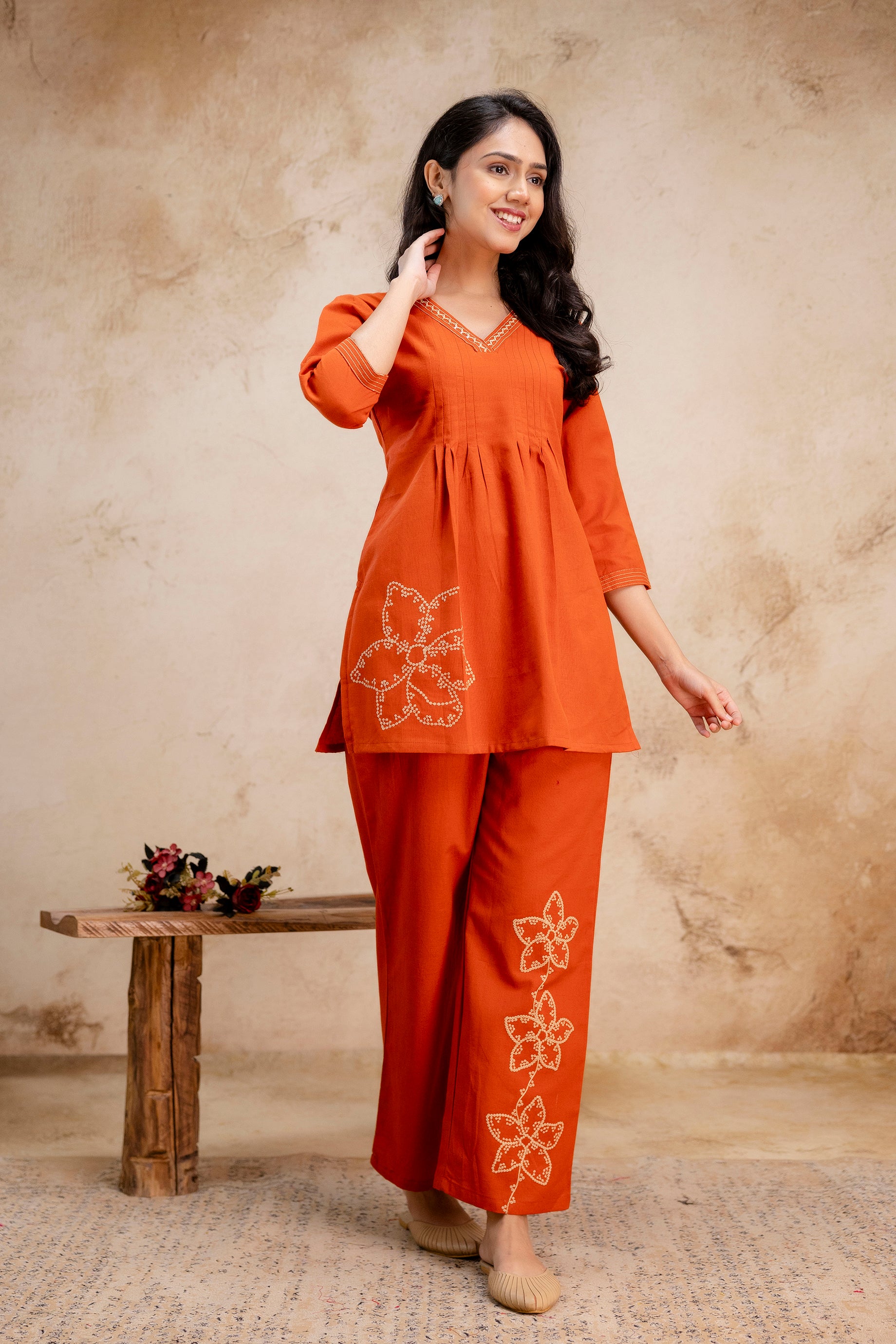 Woman in a burnt orange kurta set with white floral embroidery

V-neck kurta with pleated front and 3/4 sleeves

Large embroidered flower on left side of the kurta

Three floral motifs on lower pant leg

Indoor setting with soft beige background