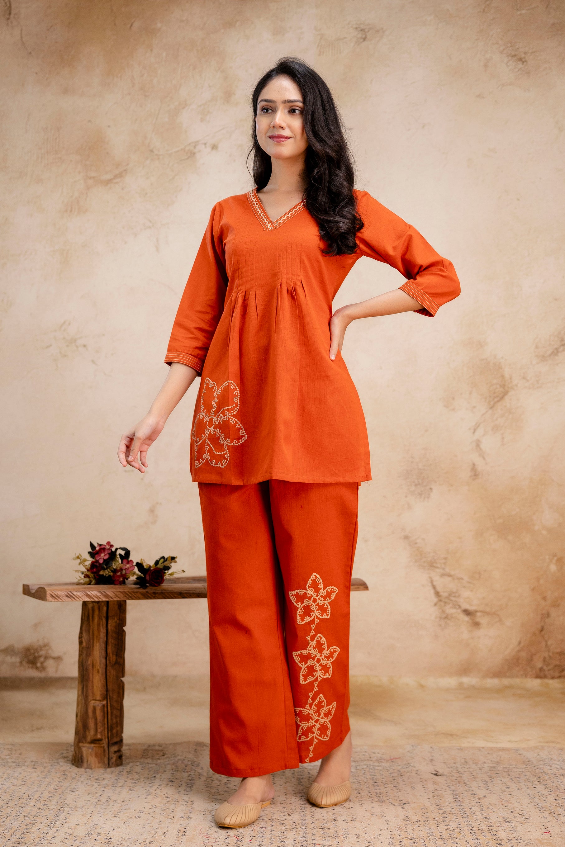 Woman wearing a burnt orange embroidered co-ord set

V-neck kurta with pleated front and floral motif on the left hip

Straight-fit pants featuring three embroidered flowers along one leg

3/4 sleeves and soft, breathable fabric

Styled in a minimal indoor setting with a rustic background
