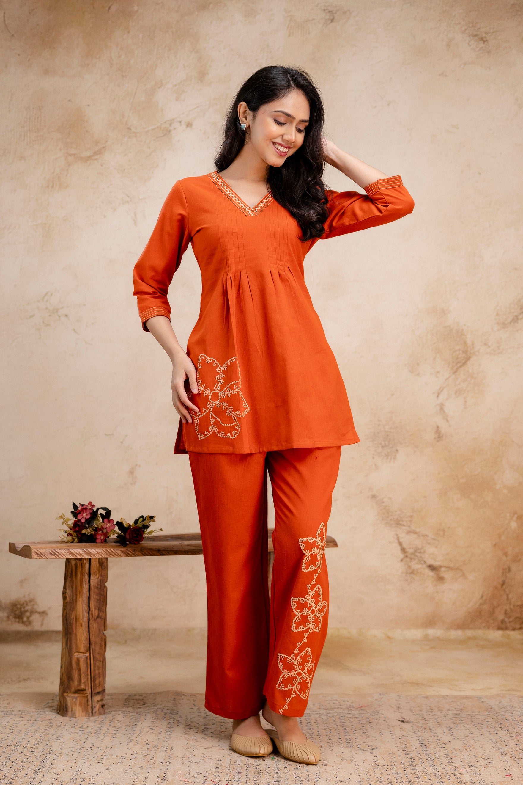 Woman wearing a vibrant rust-orange embroidered co-ord set

V-neck kurta with pleated bust and floral embroidery on one side

Full-length straight pants with three white embroidered floral motifs along the leg

3/4 sleeves with stitched detailing on cuffs and neckline

Natural indoor background with warm tones and minimal décor