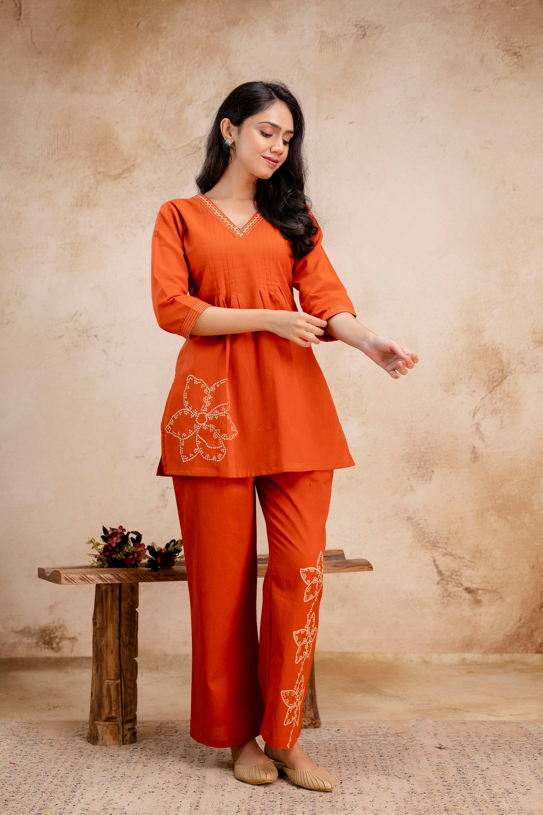 Woman wearing a burnt orange kurta set with white floral embroidery

V-neckline with embroidered detailing and front pleats

Three-quarter sleeves with stitched trim

Embroidery placed on the left pocket and lower right leg

Model adjusting her sleeve while standing on a neutral indoor background