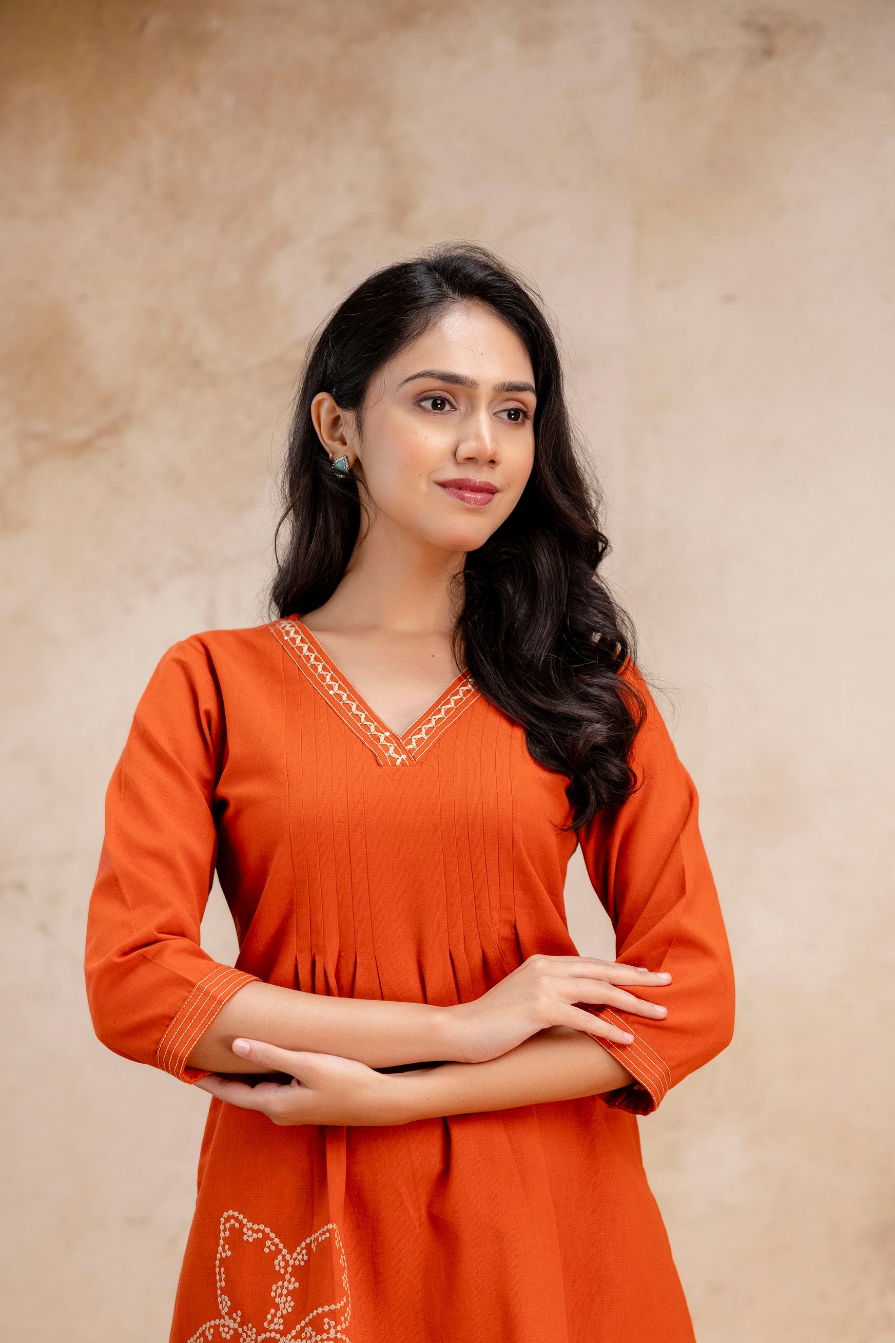 Close-up of a woman wearing a rust-orange V-neck kurta

Fine pleated detailing on the bust area for a structured fit

Embroidered neckline with white geometric pattern

3/4 sleeves with matching white stitch detail on cuffs

One visible floral embroidery on the lower left side of the kurta

Natural background with soft beige tones enhancing the warm outfit color