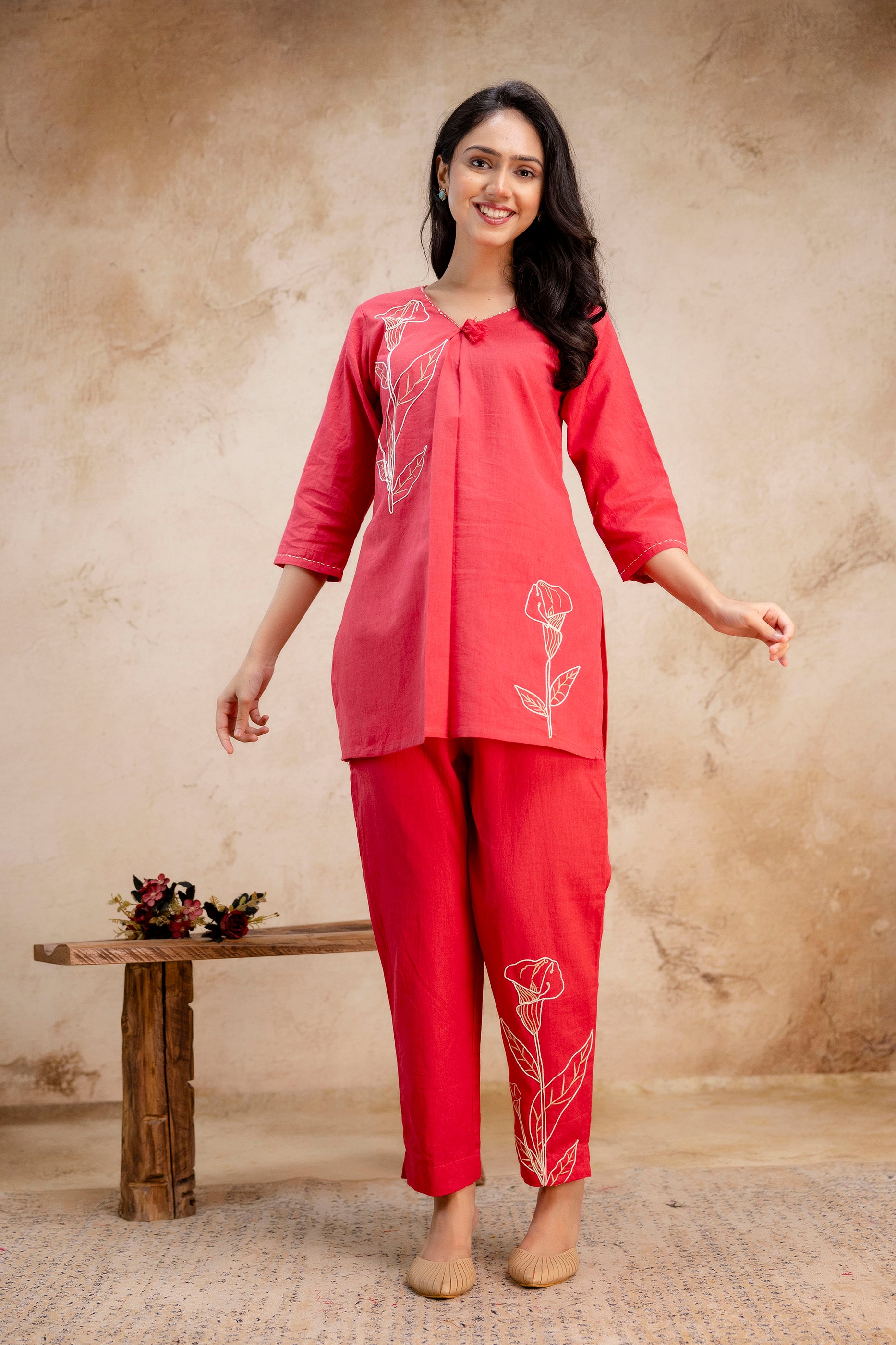 Woman wearing a vibrant red kurti and pant set with white floral embroidery.

V-neck kurti with three-quarter sleeves and tassel detail.

Matching red straight pants with floral embroidery at the ankle.

Styled with neutral juttis and minimal accessories.

Ideal for casual or festive ethnic wear.