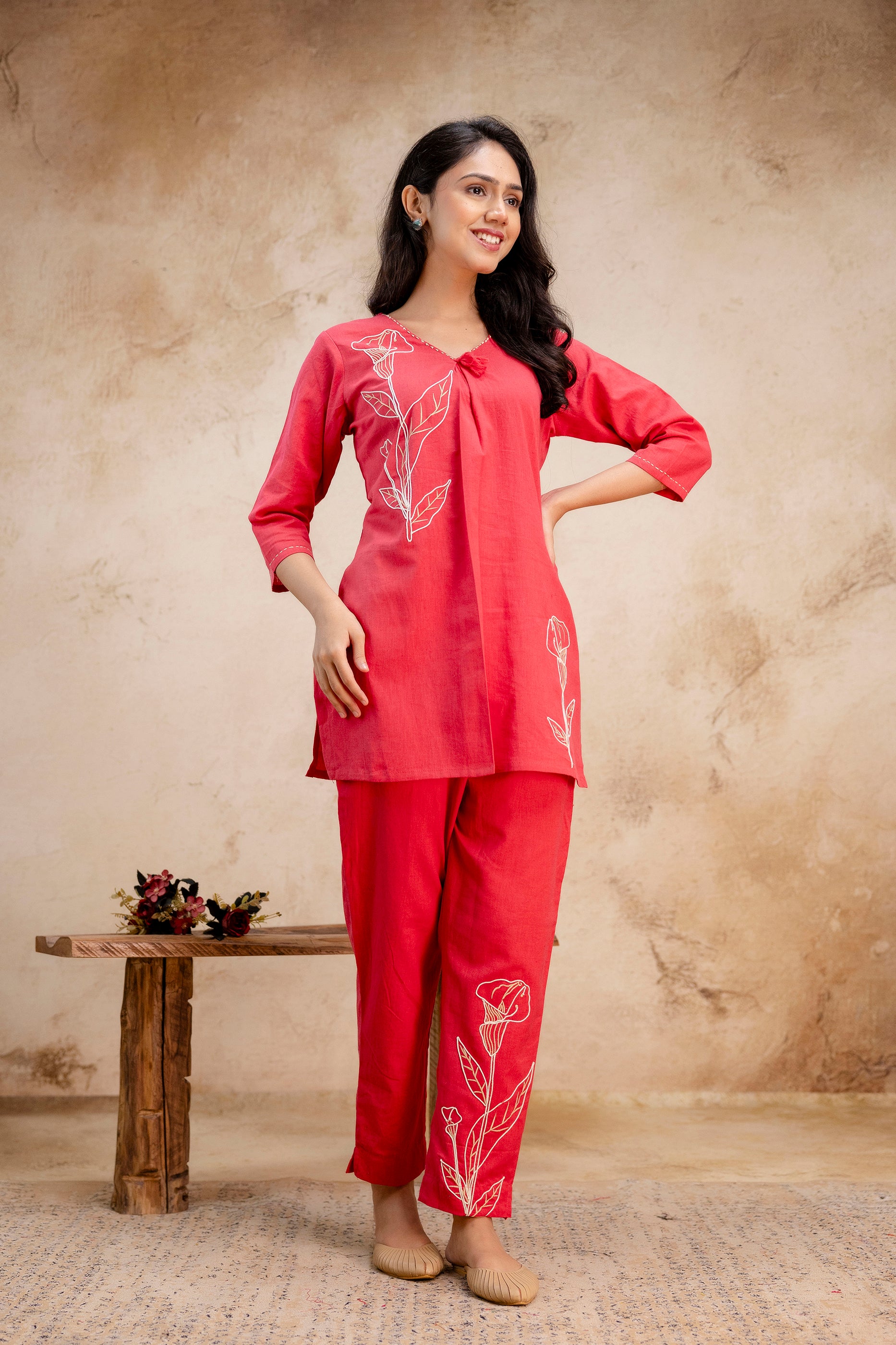 Woman wearing a bright red cotton kurti set with white floral embroidery.

V-neckline with a gathered pleat and decorative tassel detail.

Features 3/4th sleeves and straight hem for a classic silhouette.

Matching straight pants with coordinated floral embroidery on one leg.

Styled with beige juttis and minimal accessories for a chic ethnic look.

Ideal outfit for casual daywear, festive events, or ethnic office wear.