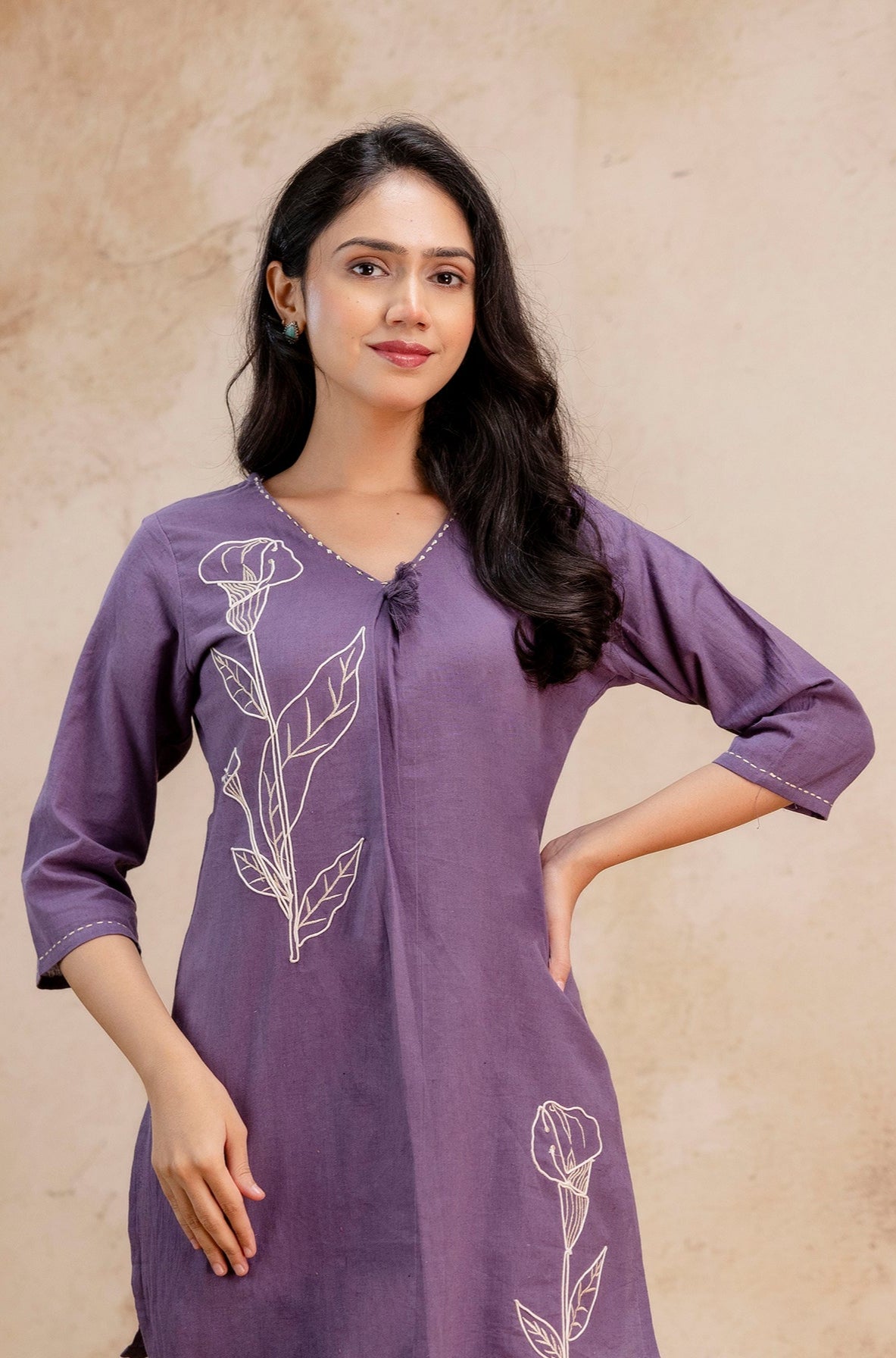 Close-up view of a stylish purple kurti set featuring elegant floral embroidery.

White thread-embroidered flower motifs enhance the upper chest and lower panel.

Three-quarter sleeves with fine detailing at the hem offer a refined finish.

V-neck design with hand-tied knot accent at the neckline for a subtle, chic touch.

Crafted in breathable cotton fabric, ideal for comfortable ethnic daywear.

Keywords: purple embroidered kurti, women’s ethnic outfit close-up, cotton kurti with floral motifs, elegant In