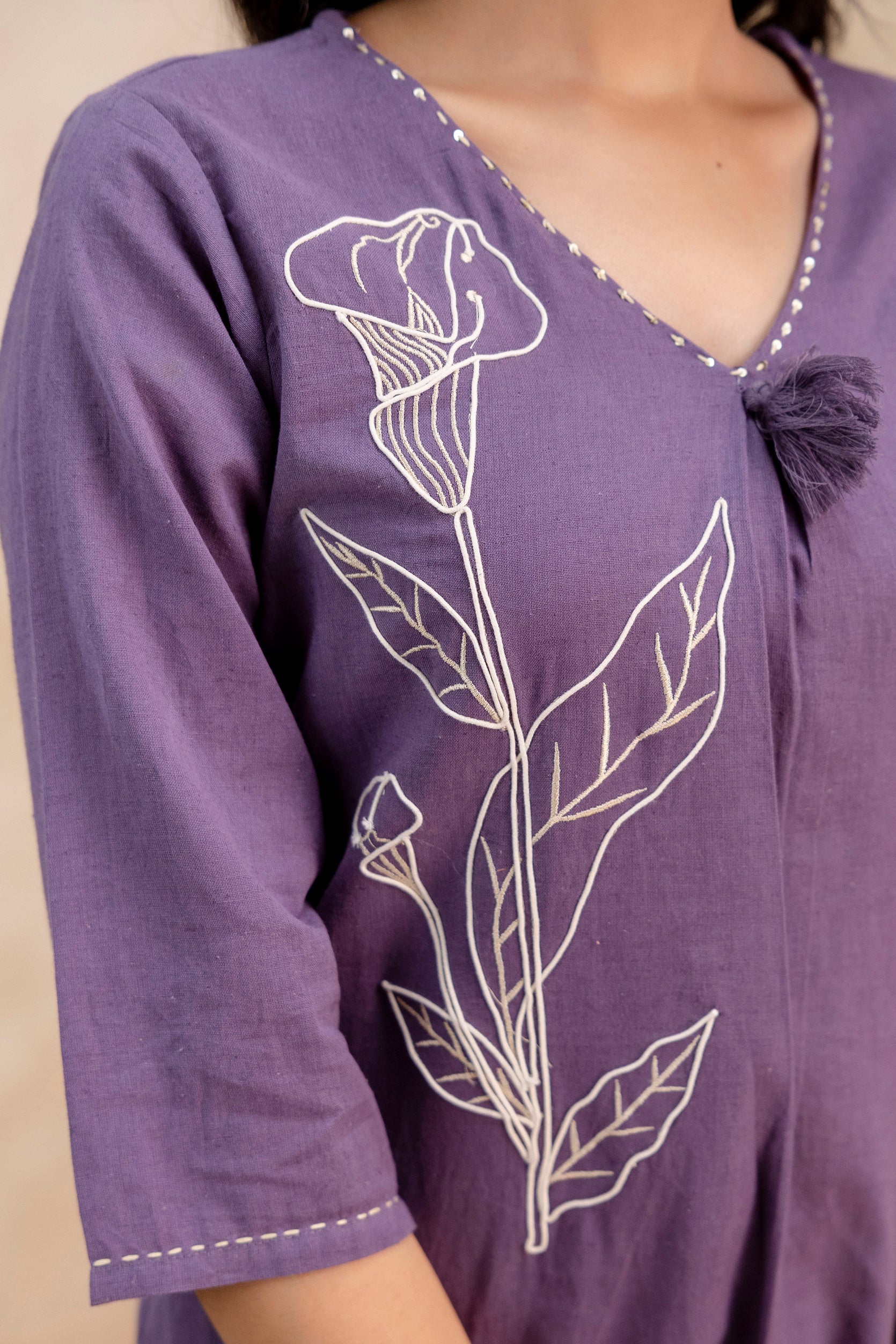 Zoomed-in view of purple kurti sleeve and chest area highlighting intricate embroidery.

Features a white floral outline embroidery with detailed leaf patterns on the front panel.

V-neckline with mirror-like stitched border and a soft tassel knot adds ethnic charm.

Three-quarter sleeve with subtle hem detailing in contrast stitch.

Cotton fabric texture is clearly visible, emphasizing comfort and breathable quality.

Keywords: embroidered kurti detail, close-up floral kurti, purple Indian ethnic wear