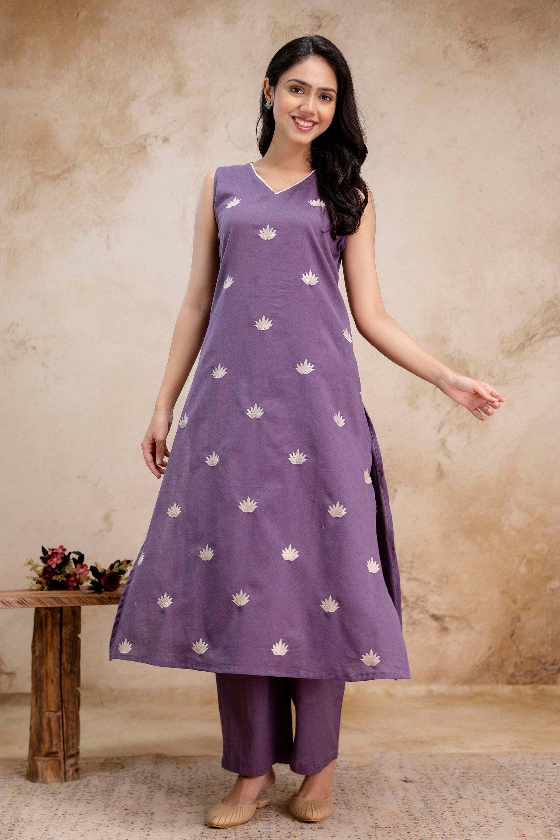 A-line kurta set in soft lavender color with sleeveless design

Fabric is embellished with subtle white lotus-like embroidered motifs arranged in a neat grid

Features a modest V-neck with light embroidery around the neckline

Paired with straight-cut matching lavender pants that fall just above the ankle

Model wears beige ethnic juttis and stands against a muted beige rustic background

Overall look is breezy, graceful, and ideal for casual or semi-formal summer occasions