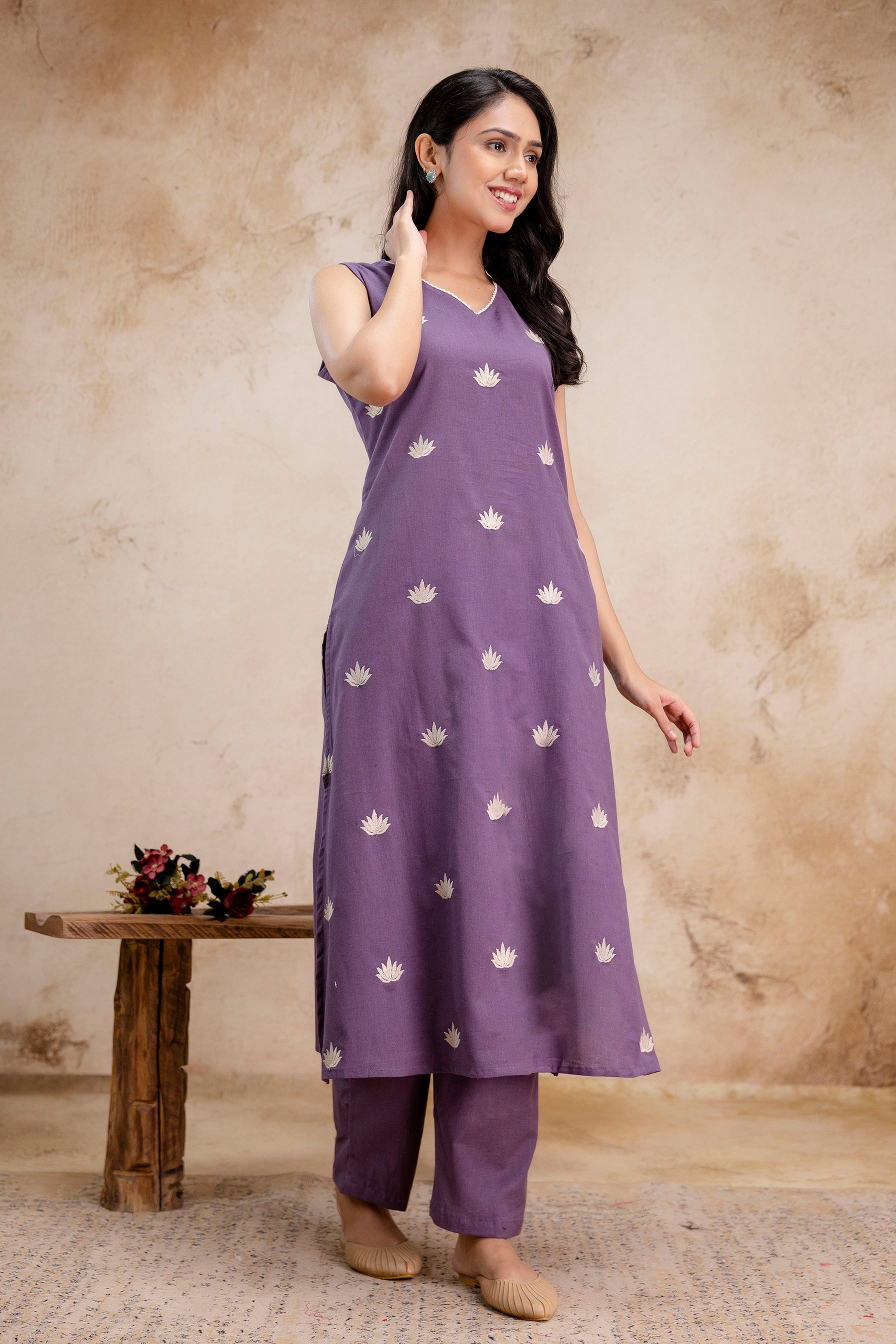 A sleeveless lavender kurta set with subtle elegance

Kurta features evenly spaced white lotus-like embroidered motifs throughout the front

Straight-cut silhouette with slight side slits for ease of movement

Paired with matching lavender ankle-length pants

Simple V-neckline with minimal embroidery enhances the clean, refined look

The model is styled with soft waves, small earrings, beige juttis, and poses against a textured beige wall

Outfit exudes a calm, summery vibe perfect for everyday ethnic wear