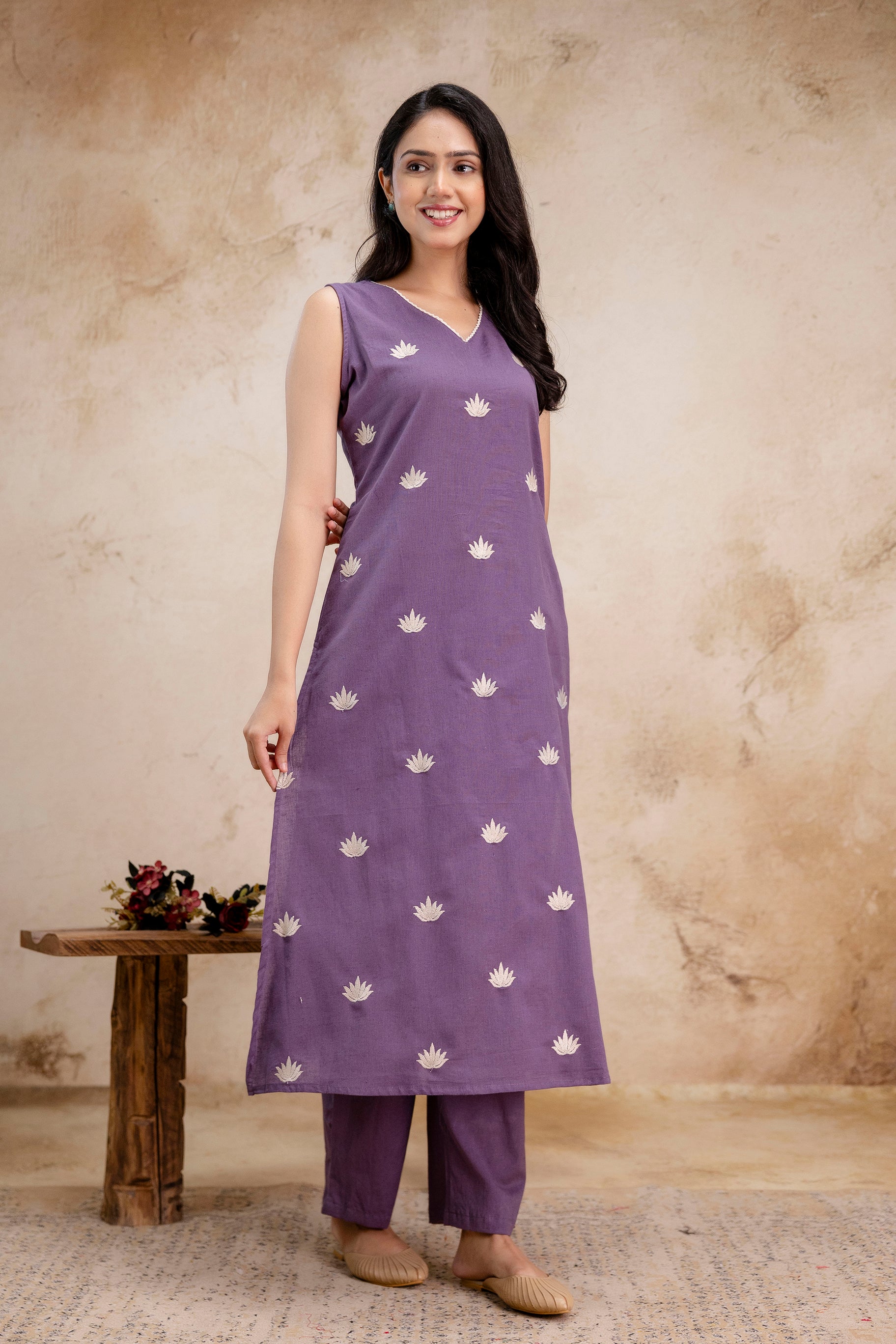 Sleeveless lavender kurta paired with straight-cut matching pants
Kurta features delicate white lotus-like embroidery in a consistent pattern across the front
Simple V-neckline with minimal stitching detail
The silhouette is clean and slightly flared, falling below the knees
Model poses with one hand resting on her waist, smiling naturally
She wears beige juttis, small earrings, and her hair is side-parted in soft waves
Background is a warm-toned textured wall, complementing the earthy calm of the outfit