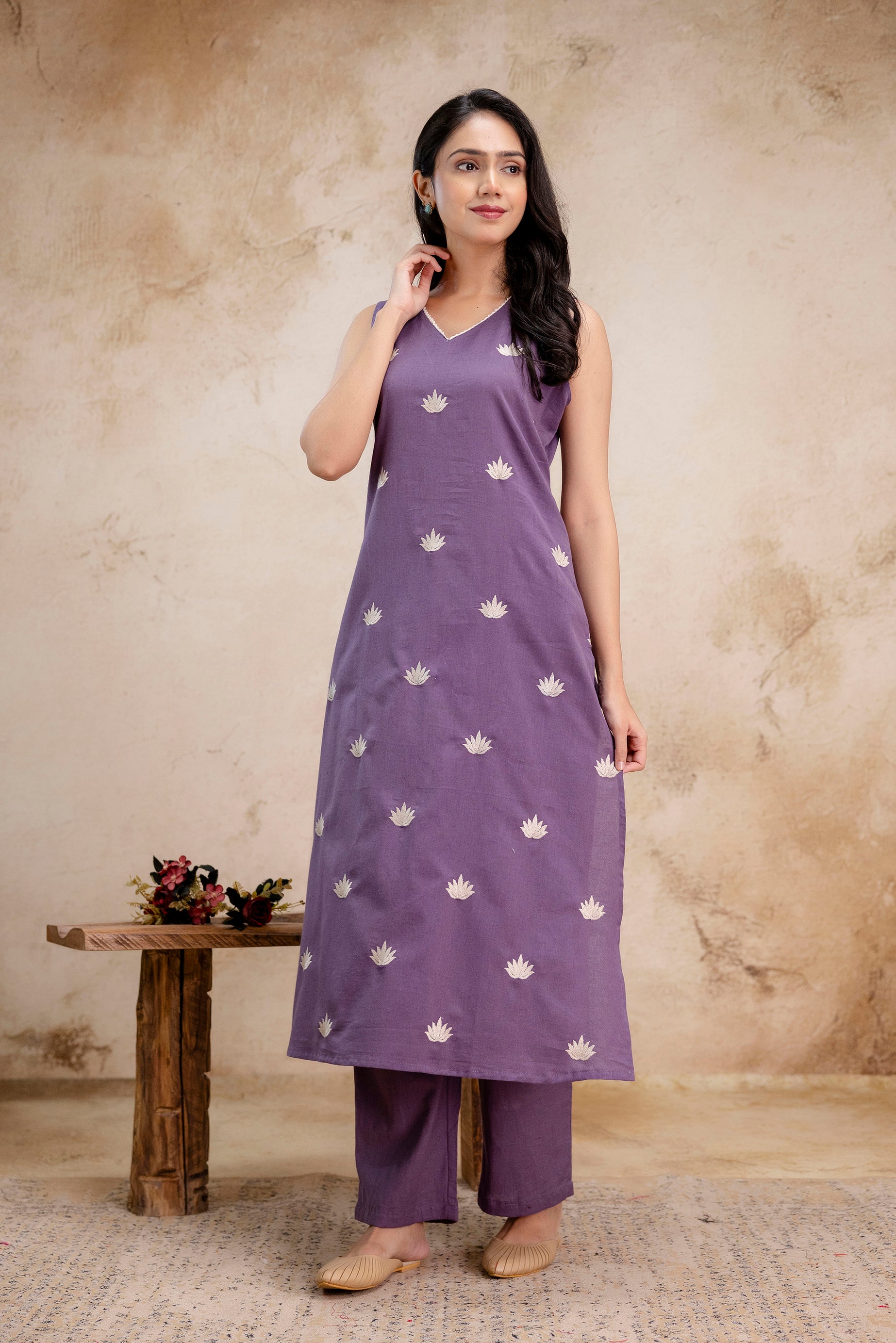 Lavender sleeveless A-line kurta with a simple V-neckline
All-over delicate white lotus flower embroidery, spaced evenly
Kurta falls below the knees and has a straight, modest flare
Paired with solid lavender straight-fit pants matching the kurta fabric
Model stands confidently, one hand lightly touching her neck
Soft curls fall over one shoulder, accessorized minimally with earrings
Footwear is beige traditional juttis
Background features a neutral-toned textured wall, keeping the focus on the outfit