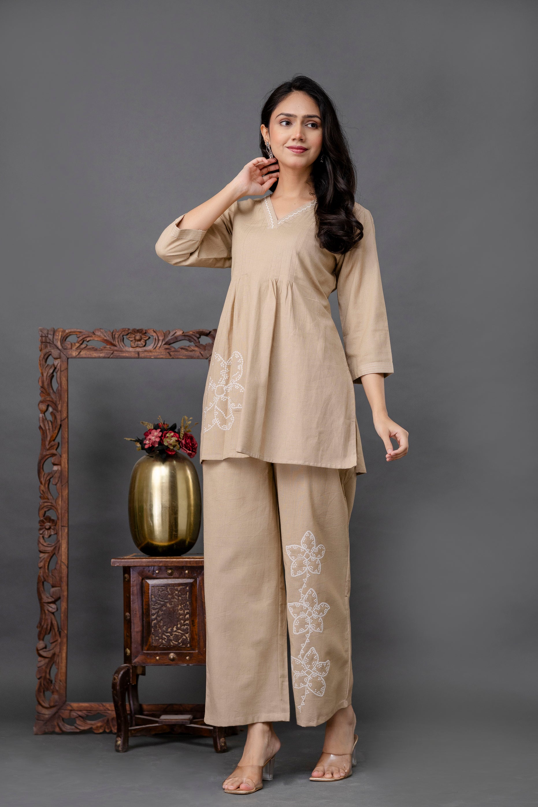 Two-piece coord set in a light beige cotton fabric.

V-neck short kurti with soft pleats starting below the bust.

Delicate white lace or thread embroidery along the neckline.

3/4th length straight sleeves for a minimal, elegant look.

Large white embroidered floral motif on one side of the kurti near the hem.

Matching straight-fit pants with ankle-length cut.

Three floral white embroidered motifs vertically aligned on one leg of the pants.

Soft structure and flowy silhouette, designed for comfort