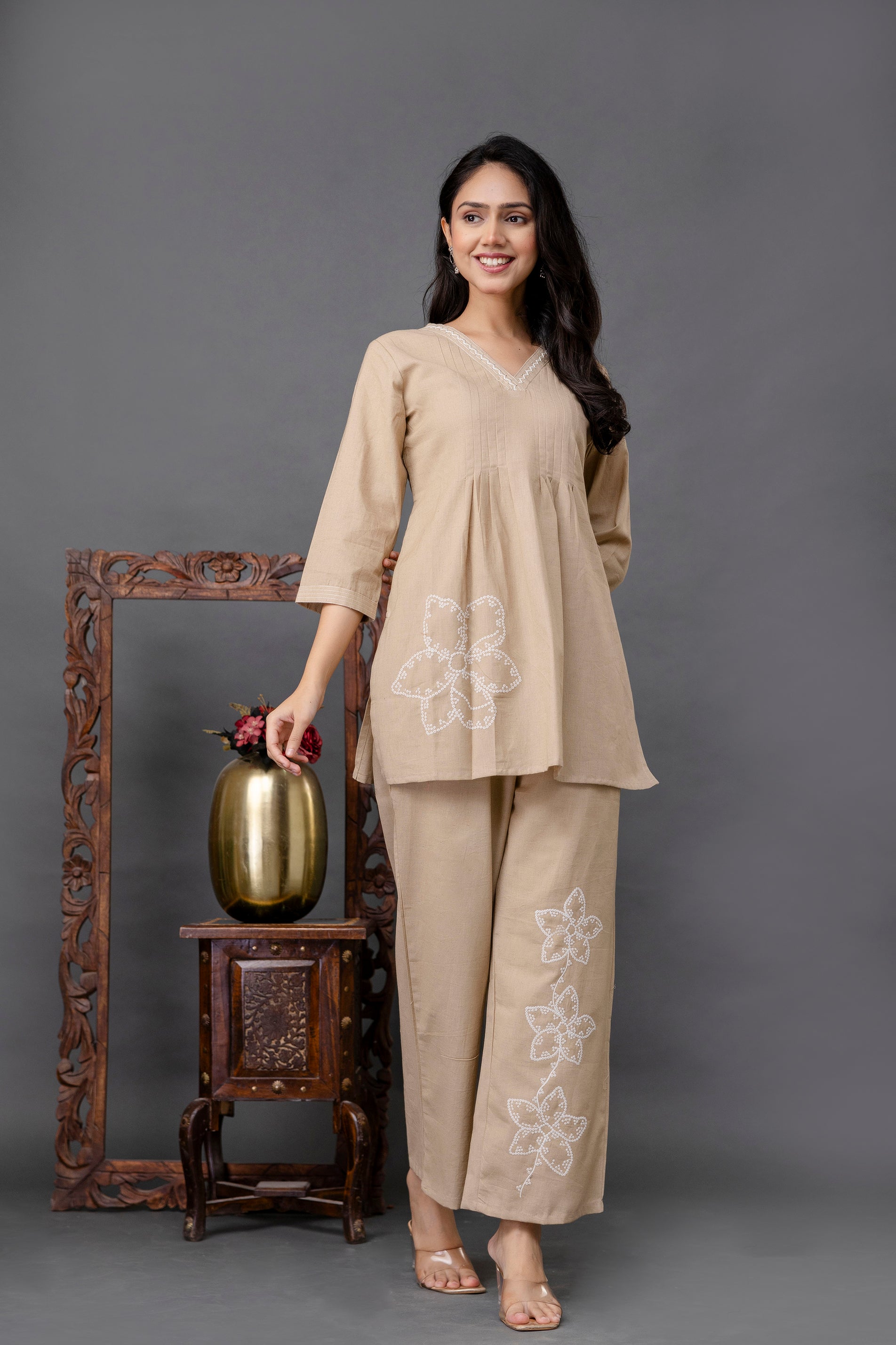 Beige cotton two-piece coord set designed for everyday elegance.

Kurti features a V-neckline bordered with fine white embroidery.

Gathered pleats below the bust for a flared, flattering shape.

3/4th straight sleeves with a clean hem.

Prominent single large white floral embroidered motif on one side of the kurti near the hip.

Straight-cut ankle-length pants in the same beige fabric.

Pants decorated with three vertically aligned white floral embroidered motifs on one leg.

Comfortable, breathable, 