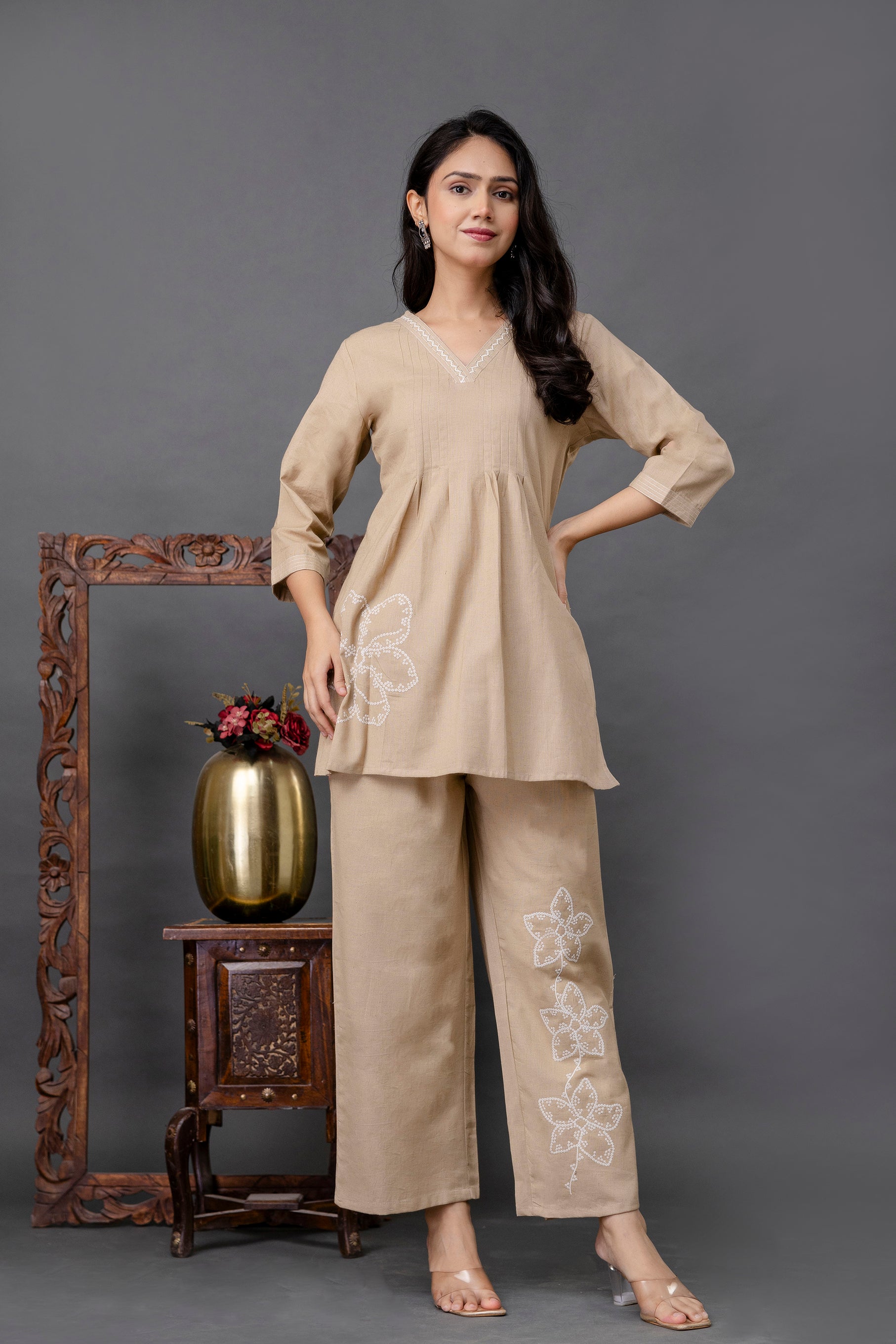 Full-length view of a coordinated beige kurti and pants set worn by a smiling female model.

The kurti features a soft V-neckline with delicate white embroidery detailing along the collar.

Subtle gathers beneath the bustline give the kurti a flared, peplum-like silhouette.

Sleeves are 3/4th length with a relaxed, straight cut.

The matching beige pants are wide-legged and adorned with large white floral embroidery motifs on one side.

Outfit fabric appears to be lightweight, likely cotton or a cotton-blen