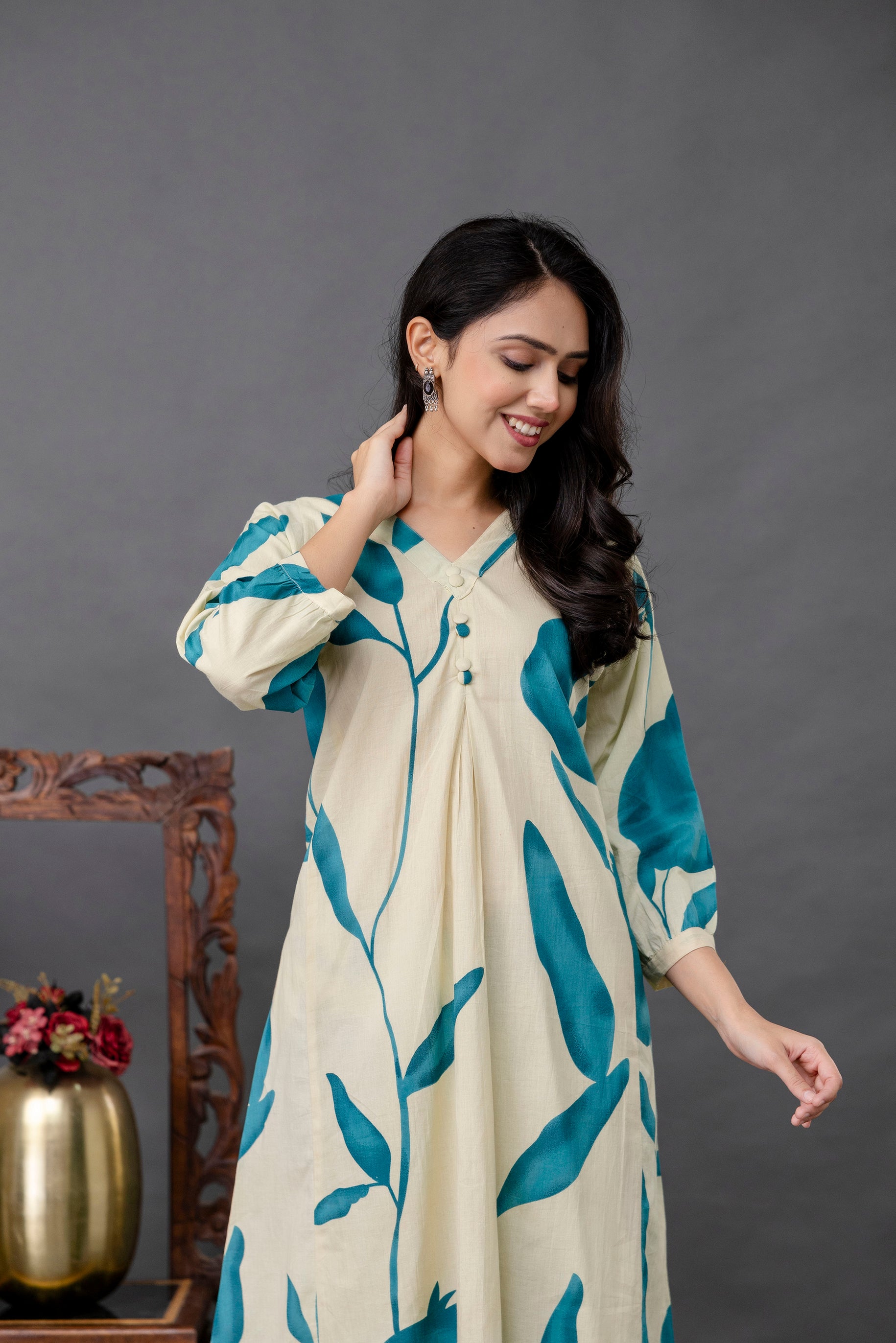 The kurti features a V-neckline with minimal button detailing and soft front pleats.

The sleeves are 3/4th length with slight gathers at the cuffs, giving a relaxed silhouette.

The teal botanical print flows vertically, enhancing the length and elegance of the kurta.

The model has long wavy hair over one shoulder and wears minimal accessories for a clean, ethnic look.

Background is a solid grey, with a traditional wooden decor and golden vase on one side, keeping focus on the outfit.
