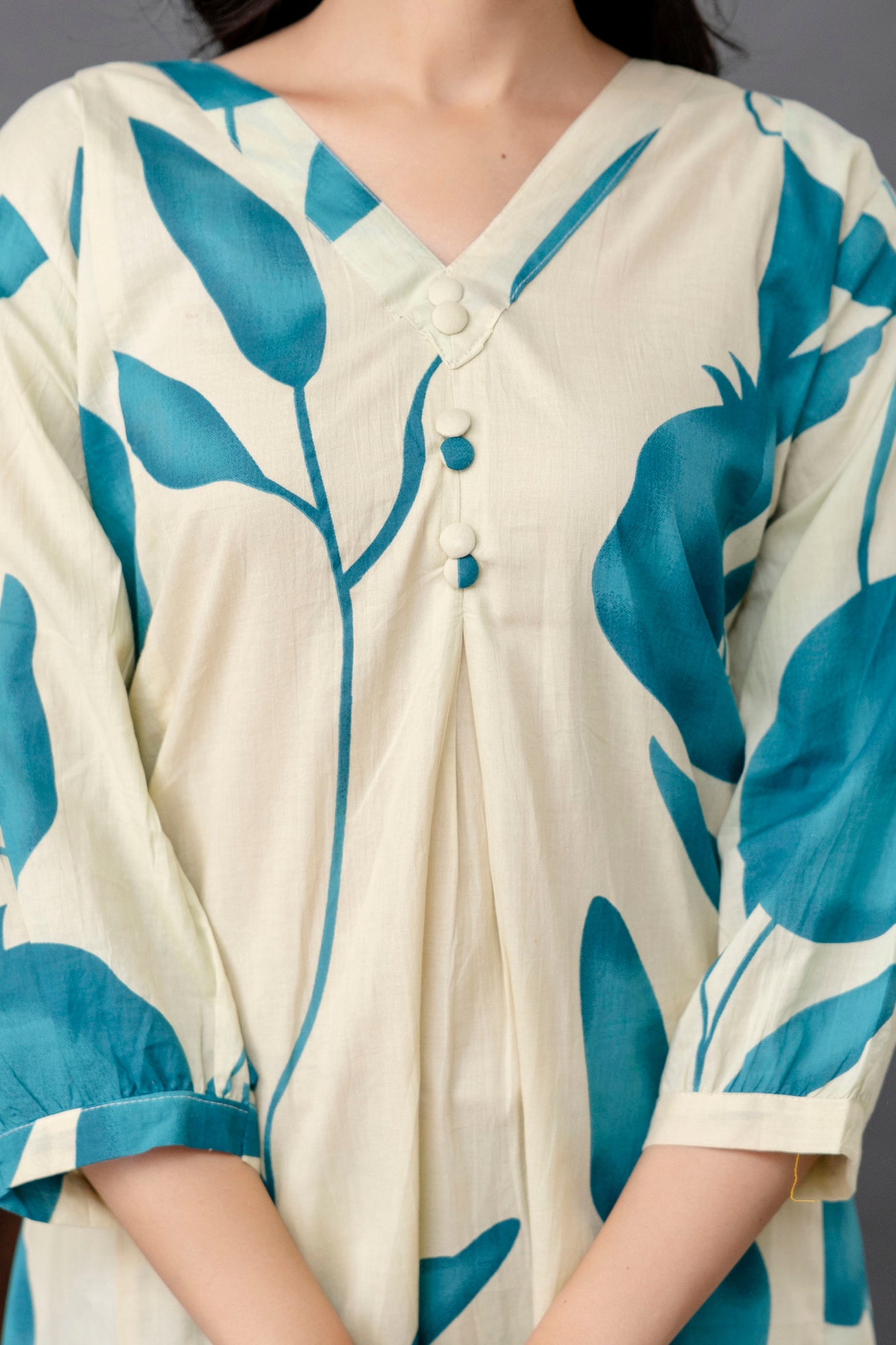 Close-up view of a cream kurti with bold teal blue botanical prints featuring abstract leaves and fruit shapes.

V-neckline accentuated with four fabric-covered buttons in matching cream and teal shades.

The pleated front adds gentle structure and flow to the garment.

3/4th sleeves are slightly puffed with a tailored hemline, echoing the teal print for cohesion.

The cotton fabric has a natural matte texture, contributing to an airy, breathable look.

The image focuses on the upper torso, showing elegant 