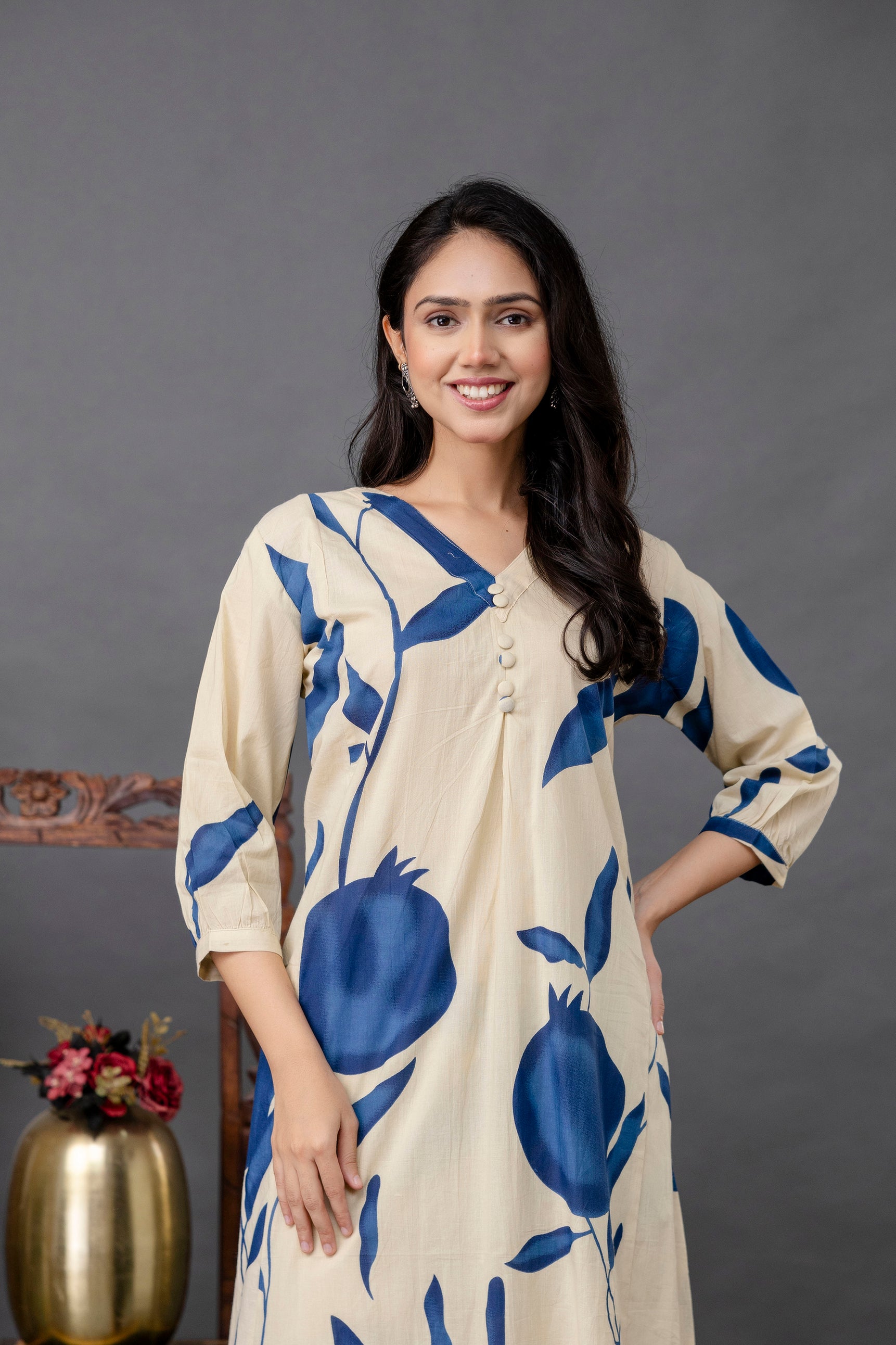 Woman wearing a beige kurta with large blue pomegranate print

Three white buttons accent the front pleated neckline

Three-quarter sleeves with bold navy motifs

Model smiling with one hand on her hip, standing against a grey background

Elegant and minimal styling with long loose hair and subtle earrings