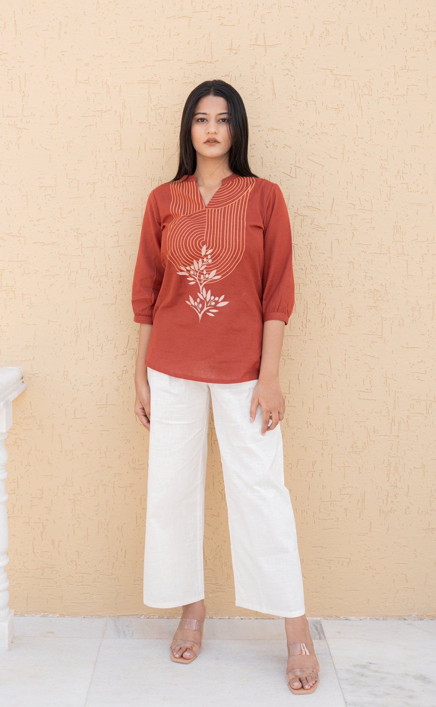 Woman wearing a rust-colored top with floral embroidery and white pants against a beige wall.