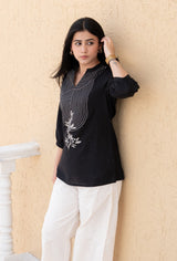 Woman wearing a black top with white patterns and white pants against a beige wall.