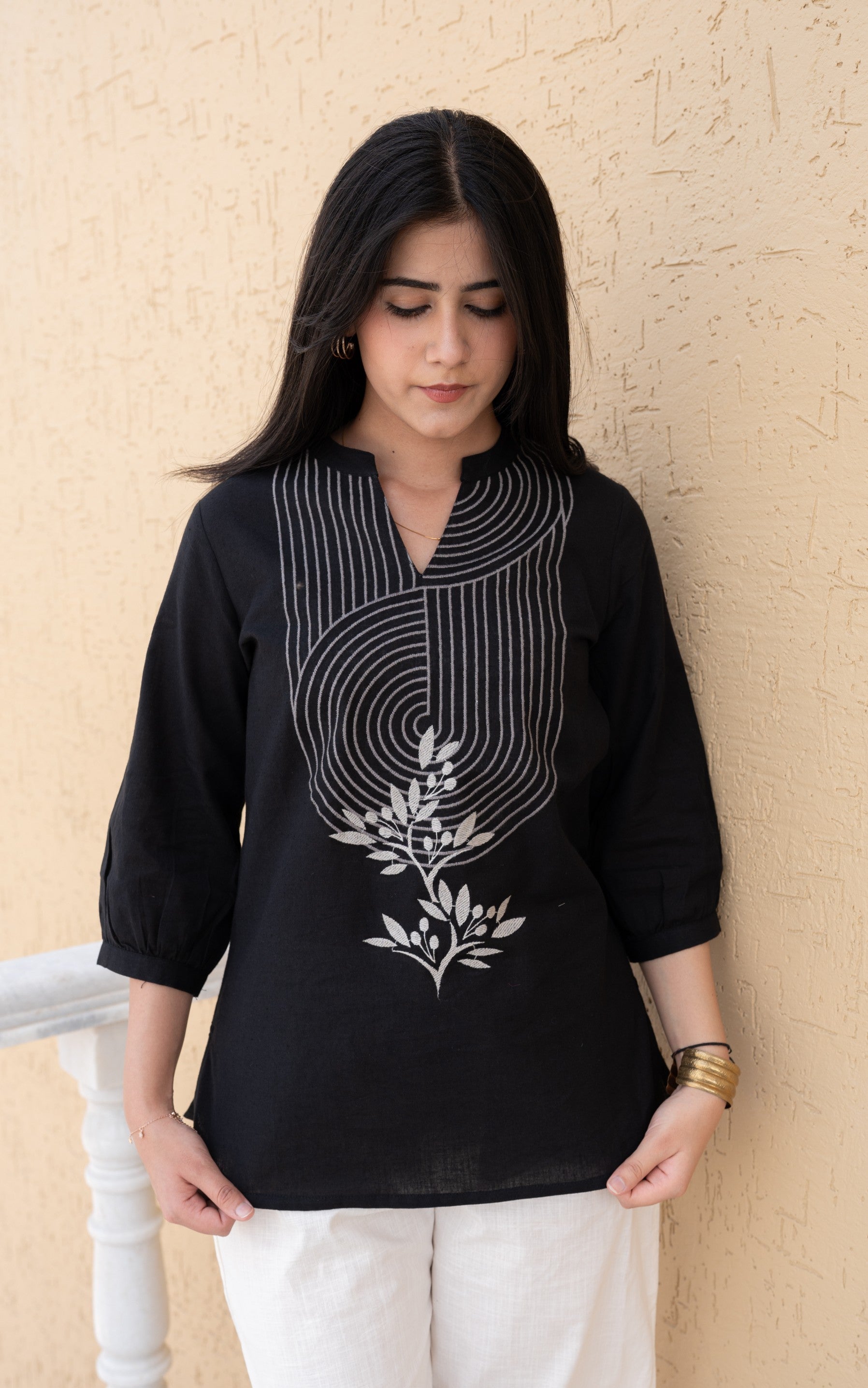 Woman wearing a black embroidered top and white pants against a beige wall.