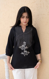 Woman wearing a black embroidered top and white pants against a beige wall.