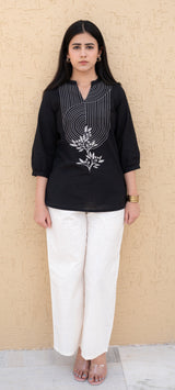 Woman wearing a black top with white floral design and white pants against a beige wall.