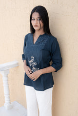 Woman wearing a dark blue top with floral embroidery and white pants against a beige wall.