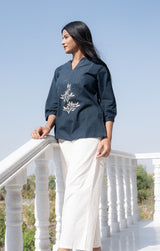 Woman wearing a dark blue embroidered top and white pants standing on a white railing with a clear sky background.