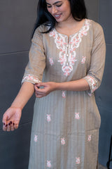 A woman wearing 'The Dusky Dupatta Set' by Tanauri, a long, light brown/khaki cotton kurti and matching trousers with delicate pink and white floral embroidery on the neckline, and holding a textured, dusty brown dupatta.