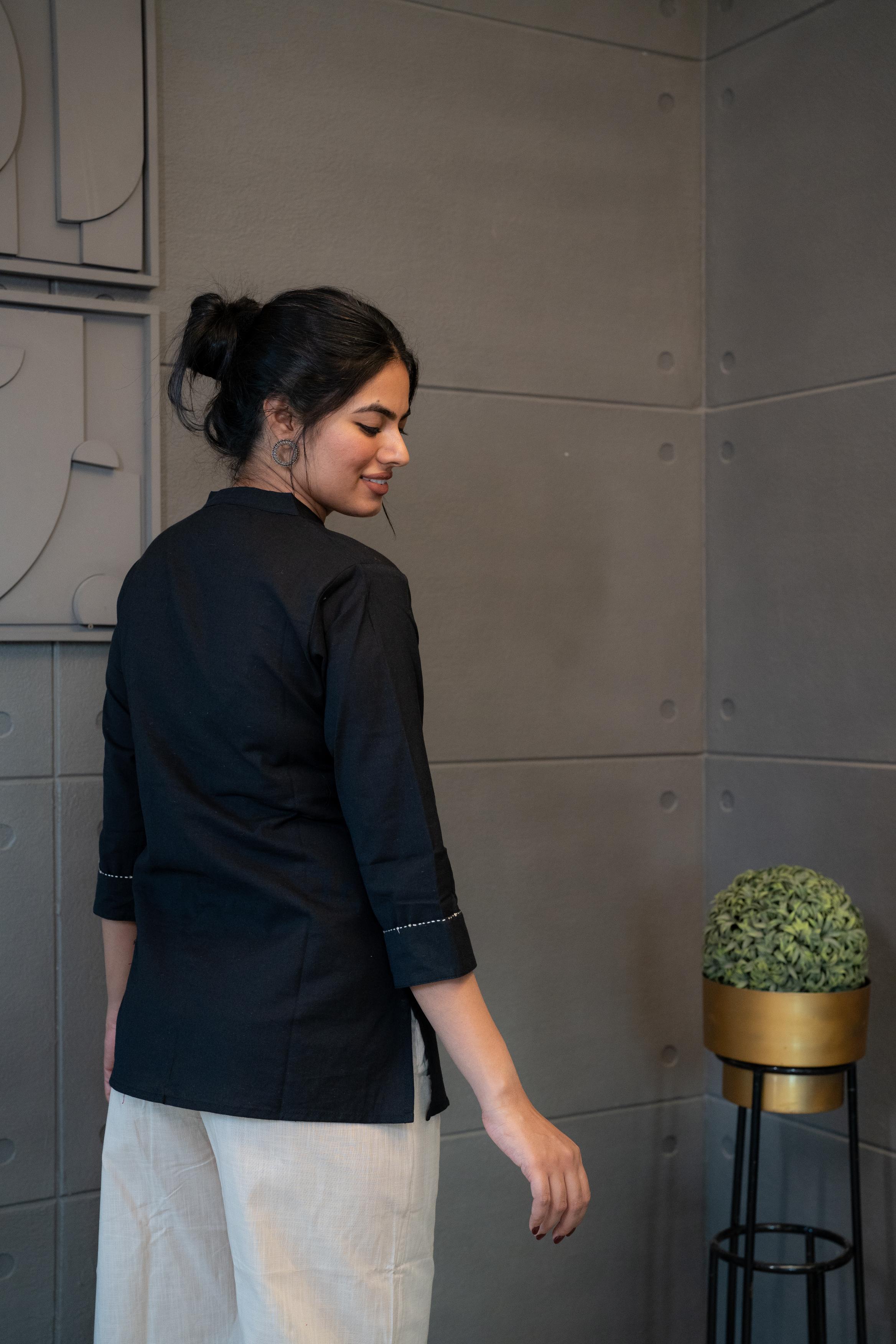 Tanauri Black Floral Short Kurti/Top, folded in clear packaging, showcasing a black cotton tunic with white floral and geometric embroidery/patchwork and wooden buttons on the placket