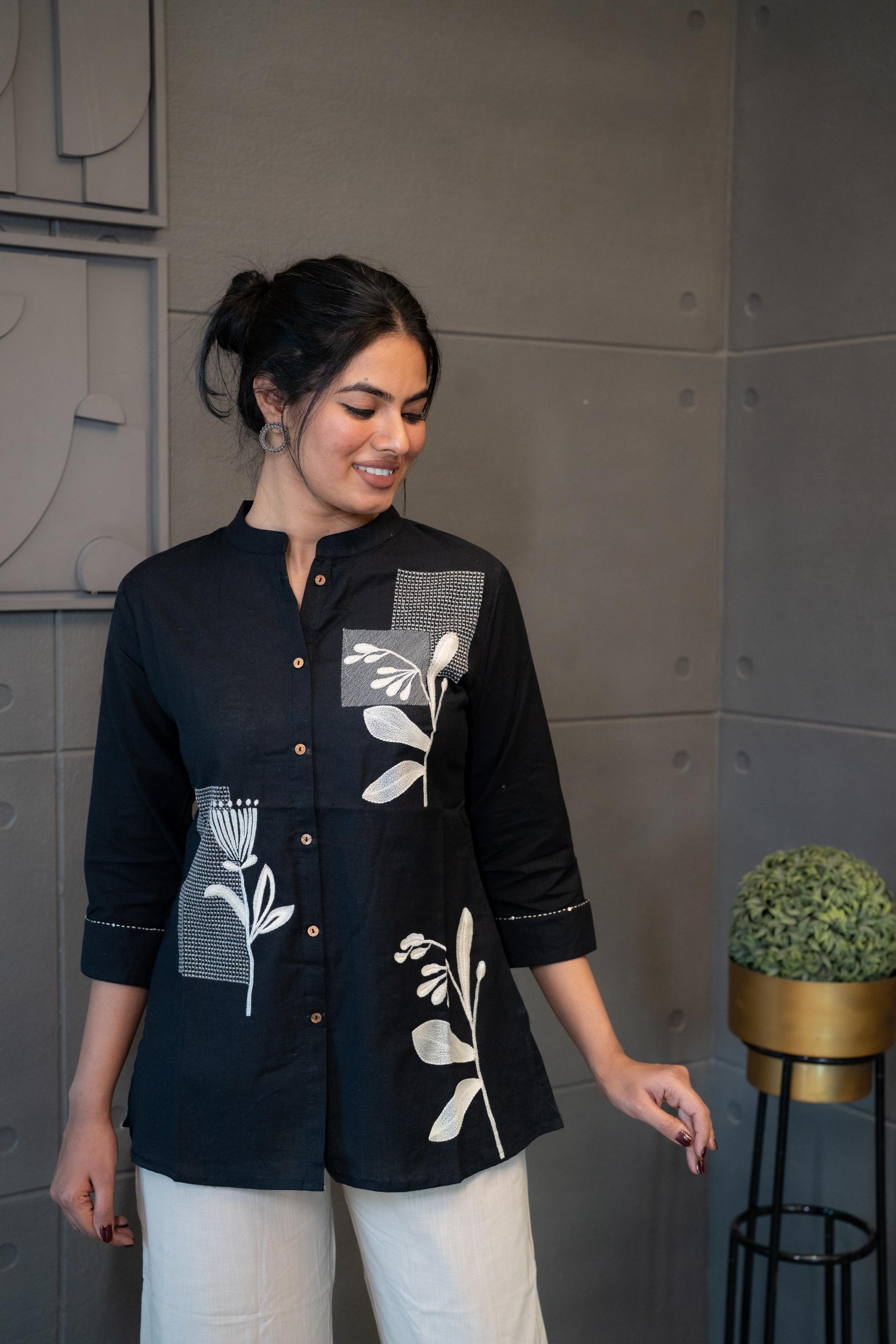 Tanauri Black Floral Top, folded in clear packaging, showcasing a black cotton tunic with white floral and geometric embroidery/patchwork and wooden buttons on the placket