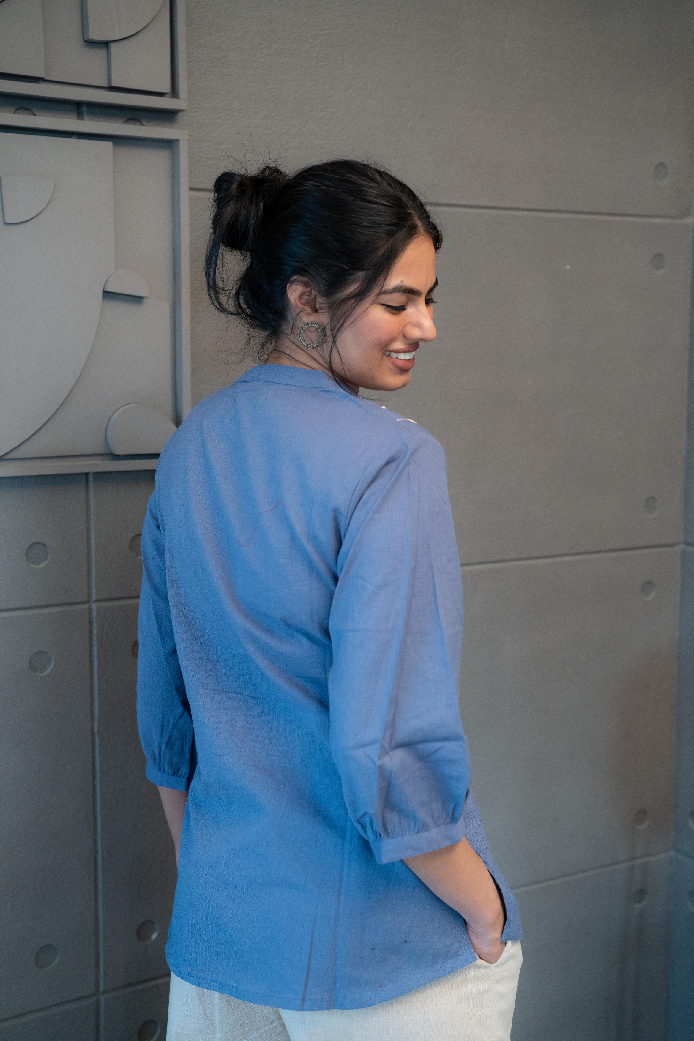 Model wearing Tanauri Blue Pop Short Kurti/Top, displaying the blue cotton short kurti with white leafy embroidery and band collar, paired with white pants for a fresh and elegant ethnic-casual look