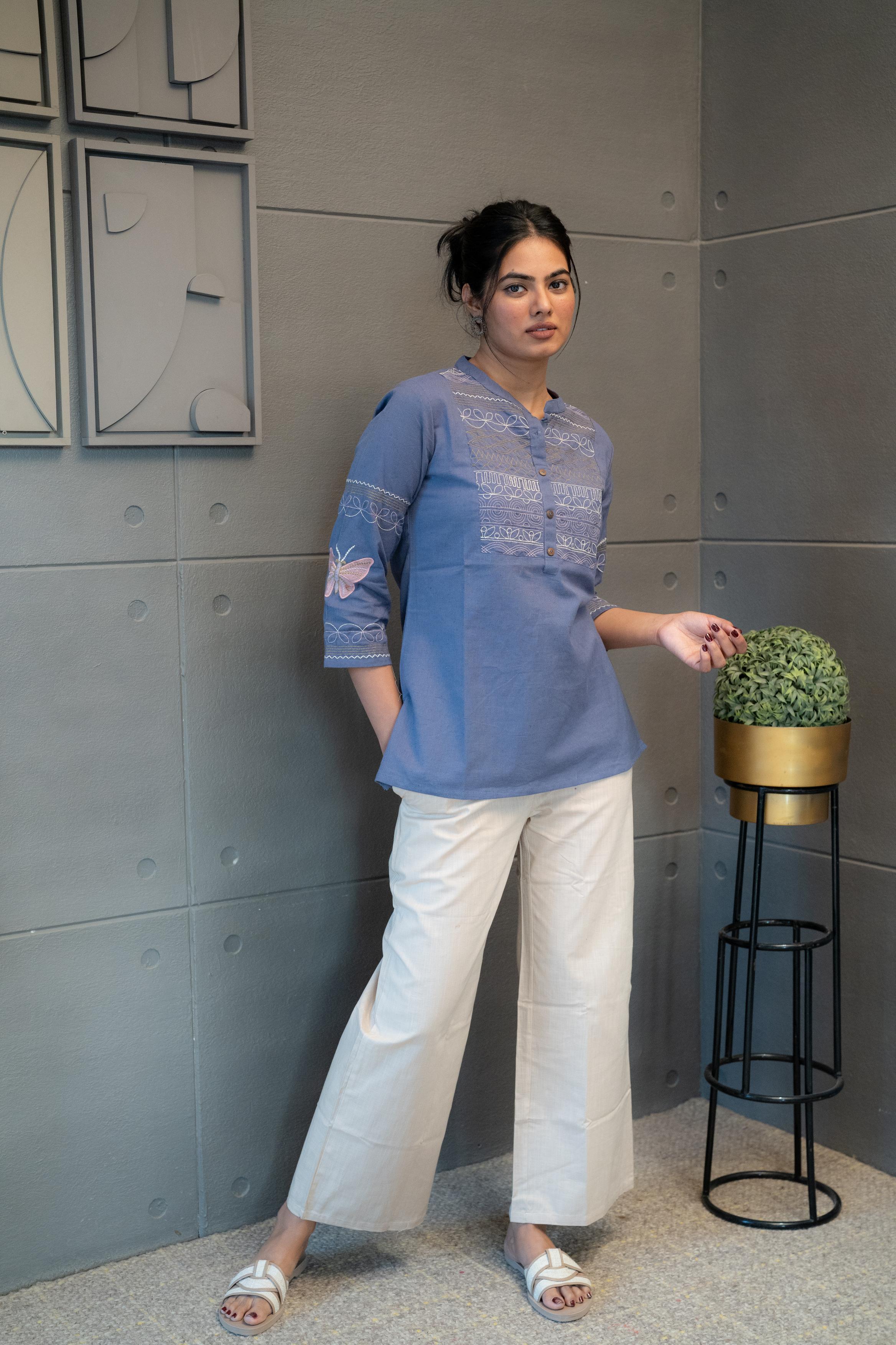 Model wearing Tanauri Orchid Short Kurti/Top, displaying the blue cotton short kurti/top with detailed embroidery and band collar, paired with white pants for an elegant ethnic-casual look