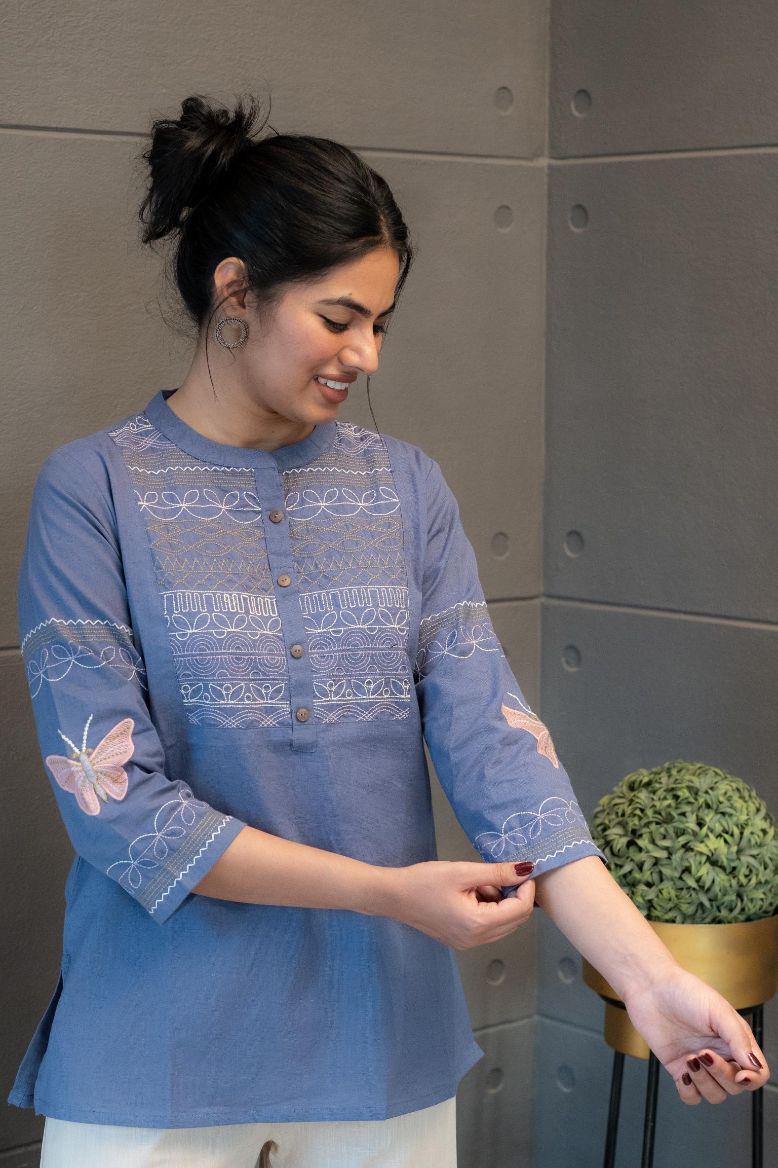 Tanauri Orchid Short Kurti/Top, folded in clear packaging, showcasing a blue cotton tunic with intricate white multi-patterned embroidery on the front and a band collar