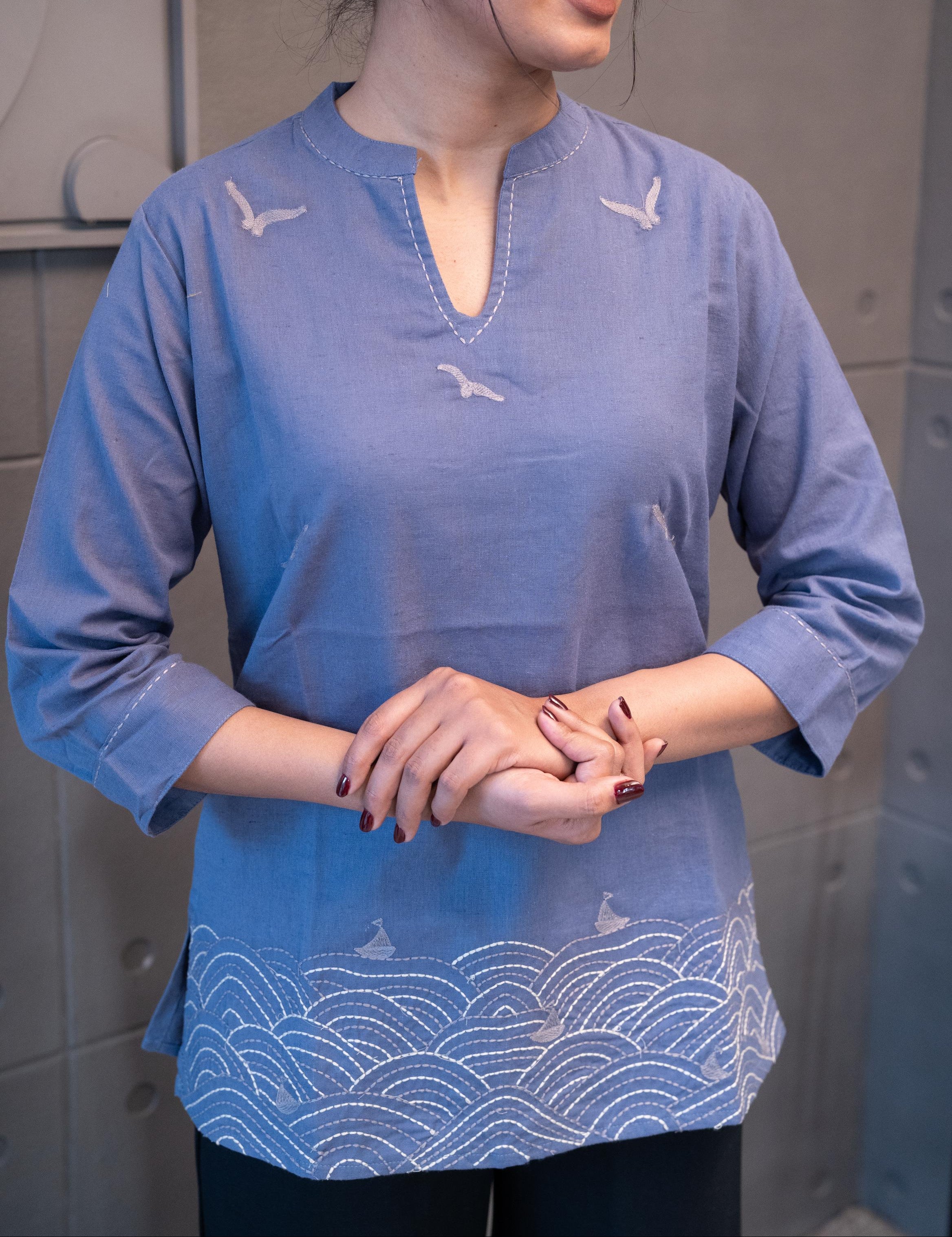 Close-up of a light grey bird motif and the matching stitching around the V-neckline on the periwinkle blue fabric of the Tanauri Grey Bird Short Kurti/Top