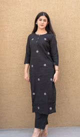 Woman wearing a black traditional outfit with silver buttons against a beige wall.