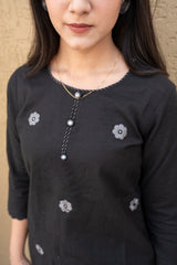 Person wearing a black top with floral embellishments against a beige wall.