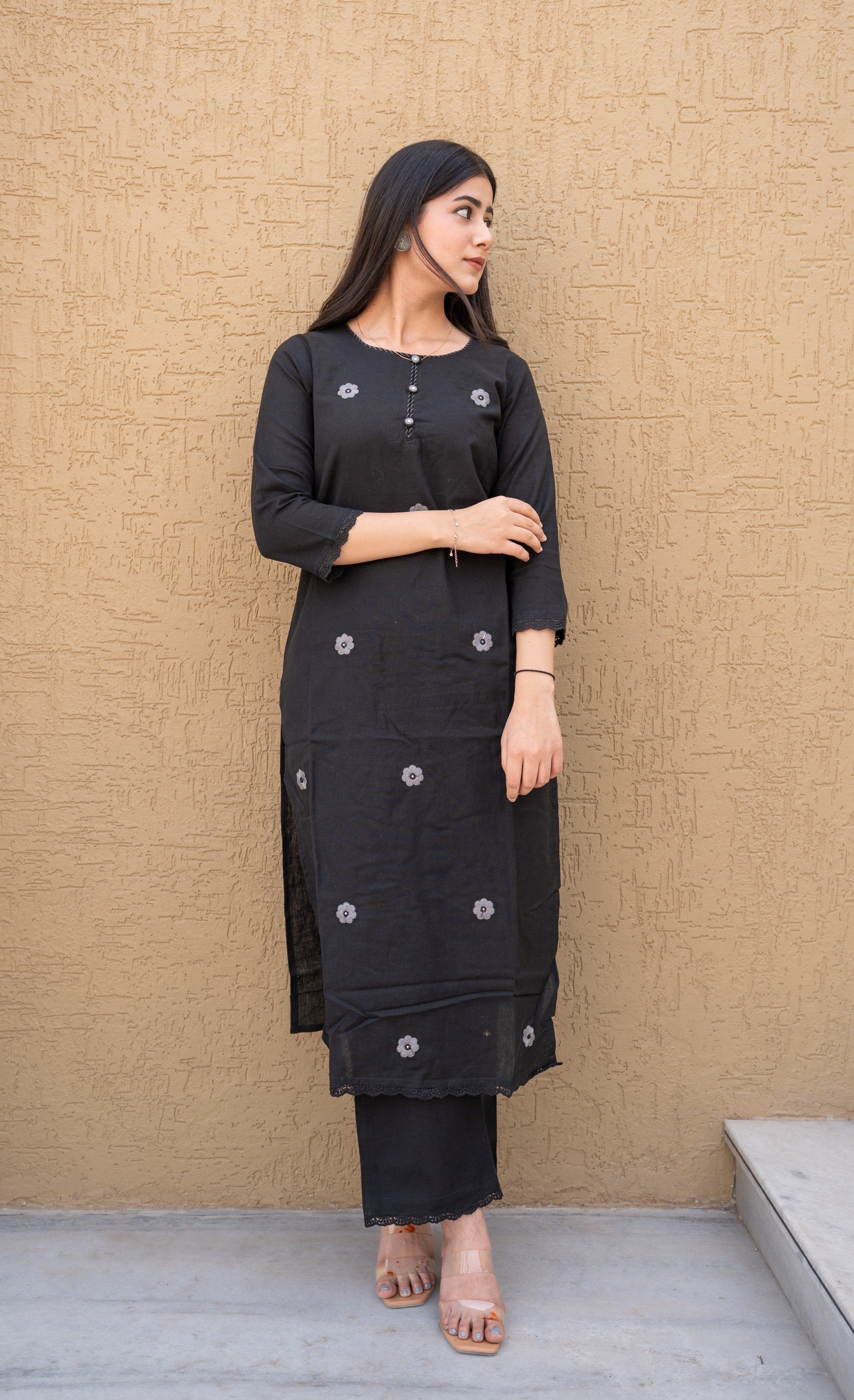 Woman wearing a black traditional outfit with floral patterns against a beige wall.