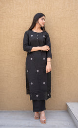 Woman wearing a black traditional outfit with floral patterns against a beige wall.