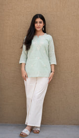 Woman wearing a light green top and white pants against a beige wall.