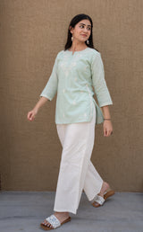 Woman wearing a light green top and white pants against a beige background