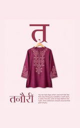 A maroon tunic top with white embroidery hanging on a wooden hanger against a pale pink background, with Hindi text and a small English quote below.